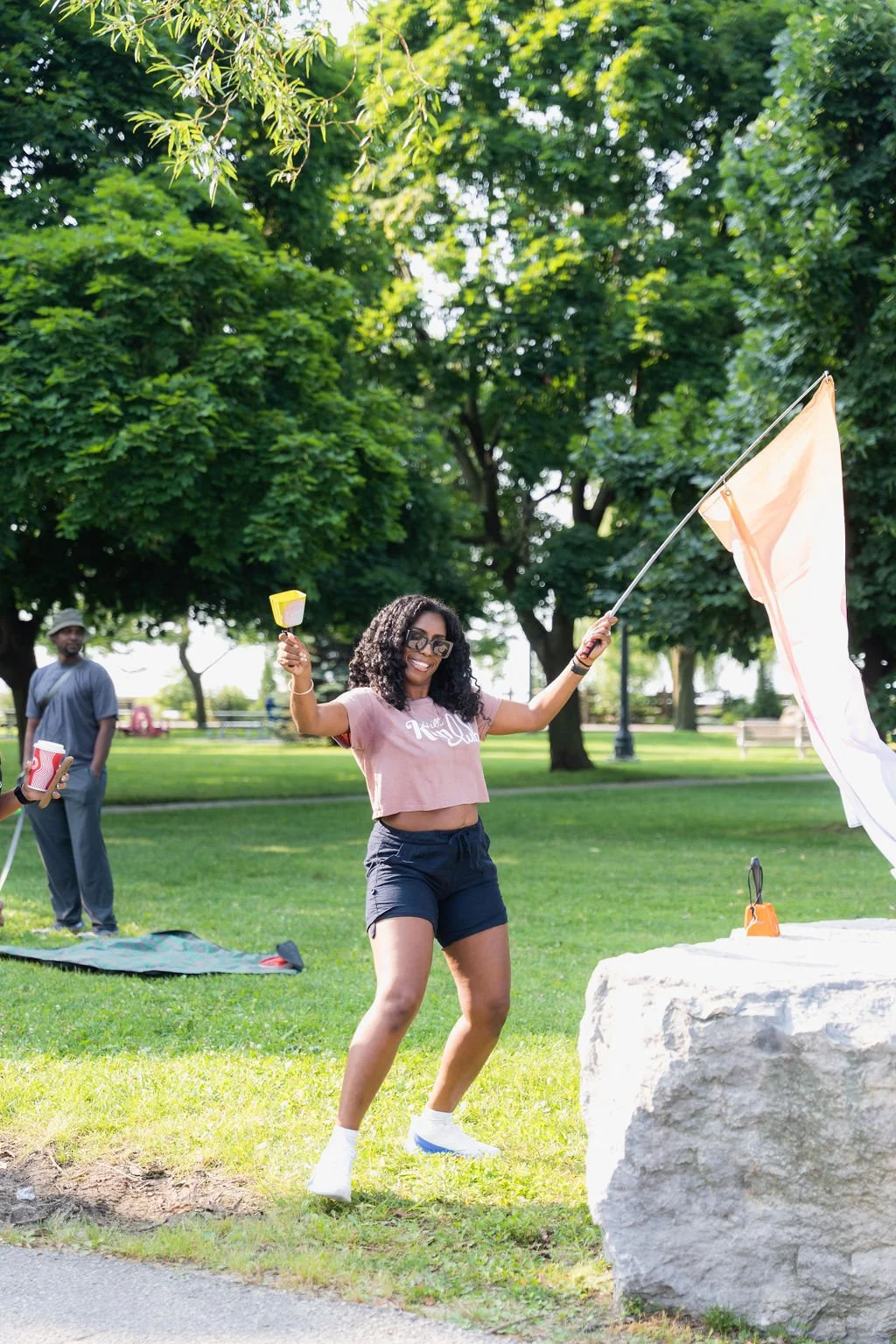 A woman with curly hair and glasses is joyfully waving a flag in a park. She is wearing a pink crop top and black shorts. Two people are in the background, one holding popcorn, with lush green trees and grass around.