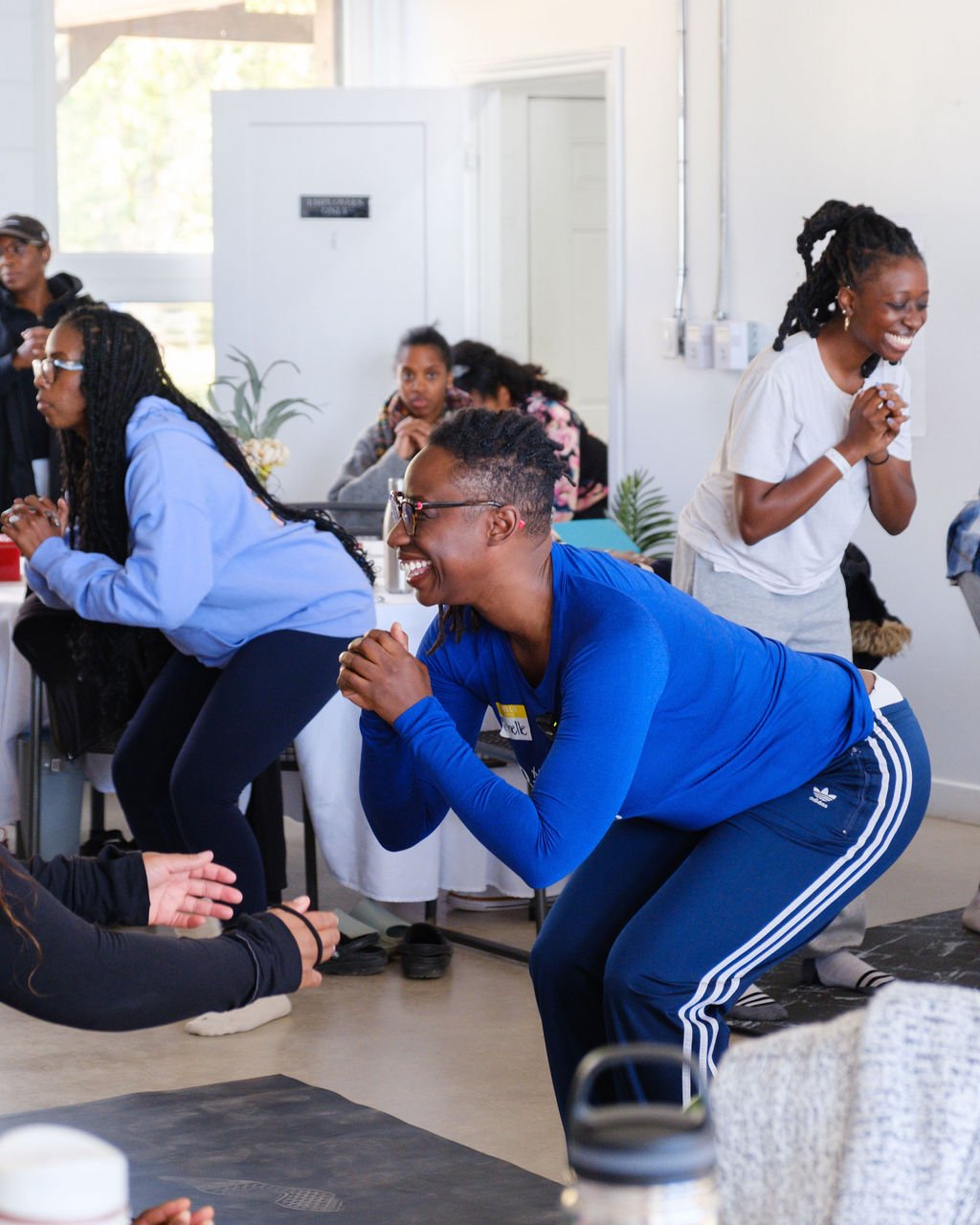 People participating in an indoor group activity or workshop, smiling and interacting cheerfully, with some kneeling and others sitting at tables.