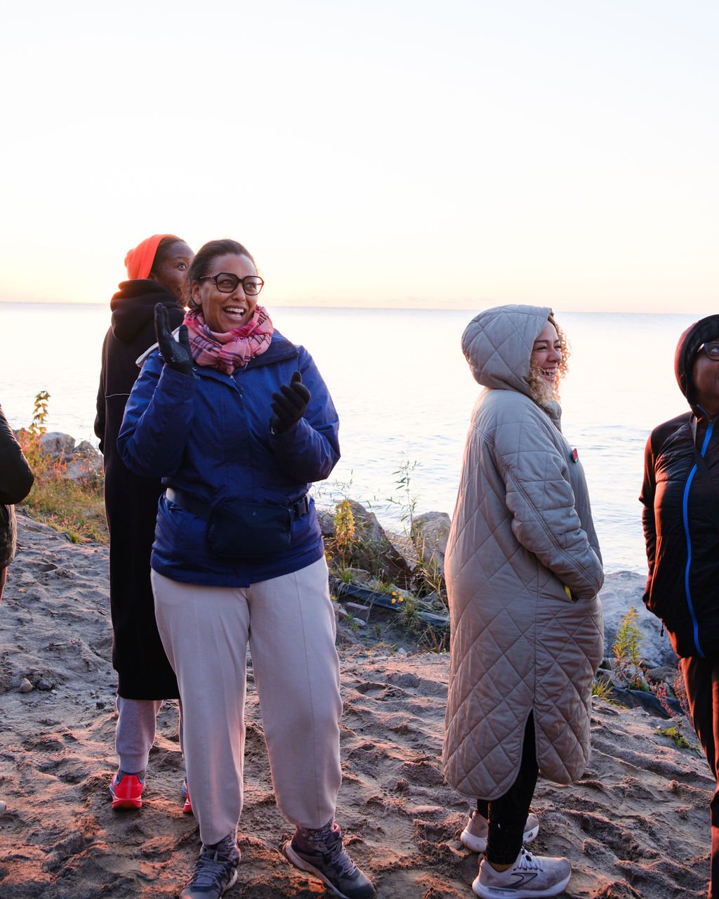 Group of people standing on a beach wearing warm jackets and hoodies, enjoying a sunset by the water.