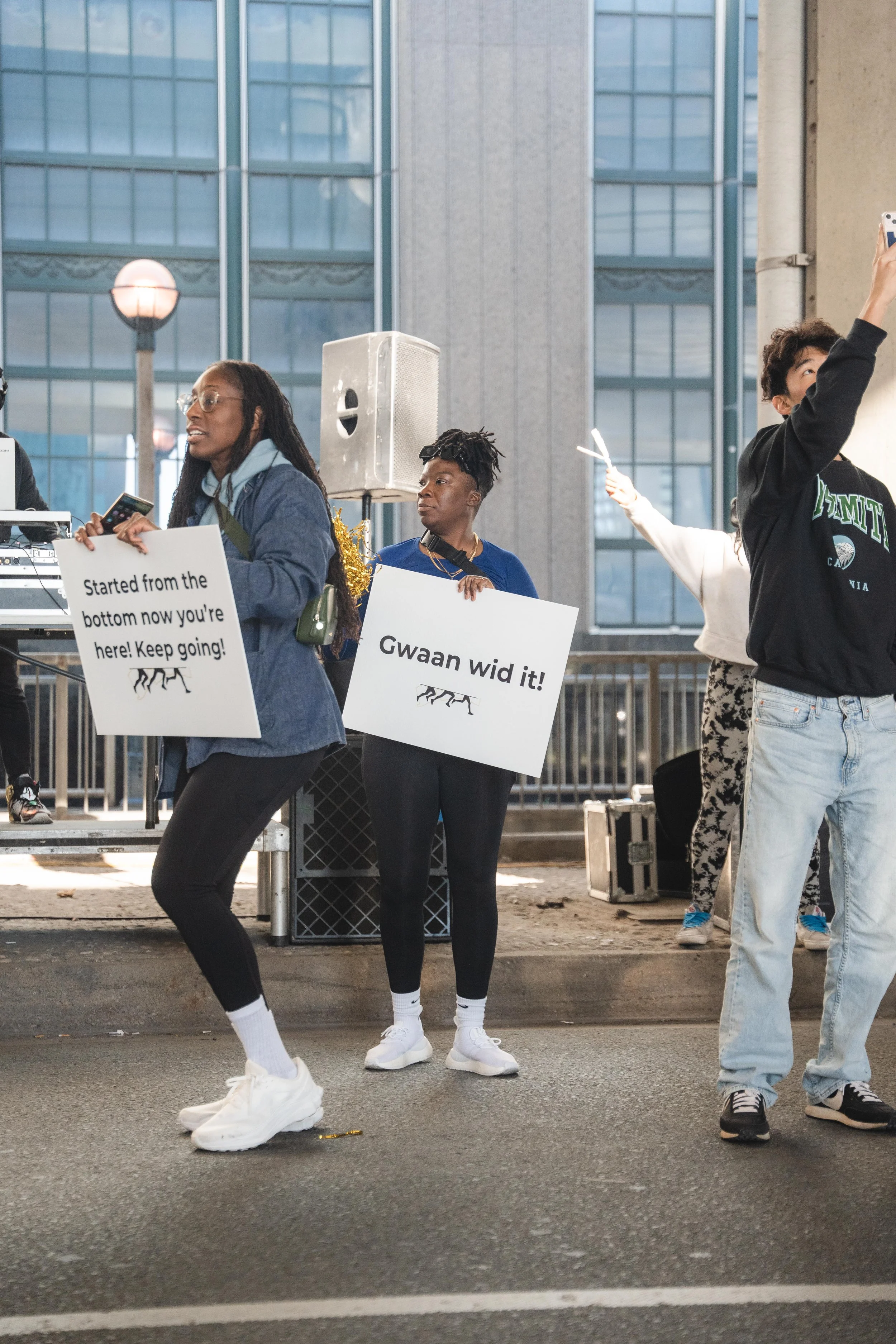 People participating in a street protest or rally, holding signs with messages. One sign says 'Started from the bottom now you're here! Keep going!' and another says 'Gwaan wid it!'