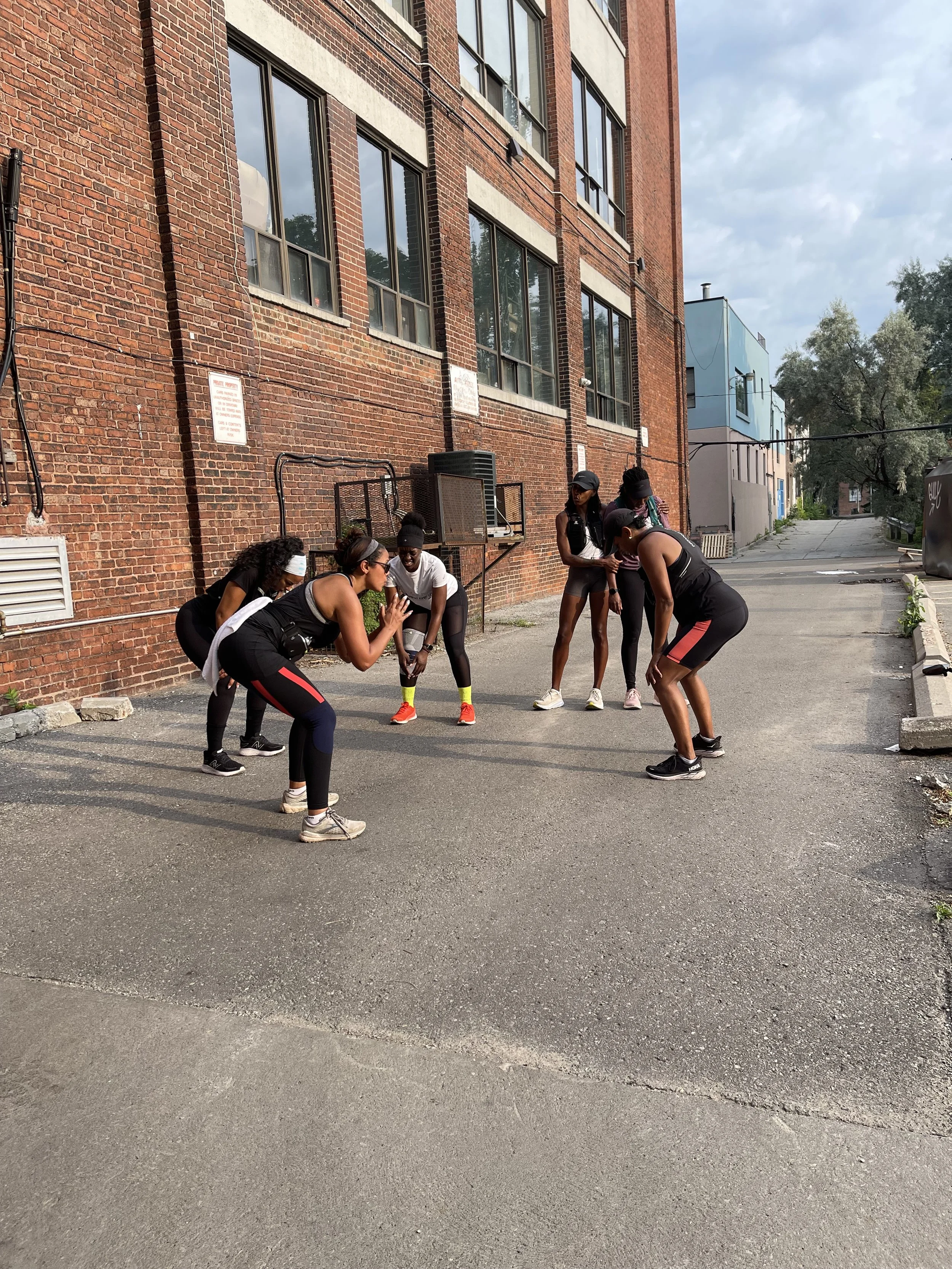 Group of six women exercising outside in an alleyway near a brick building, stretching and warming up.
