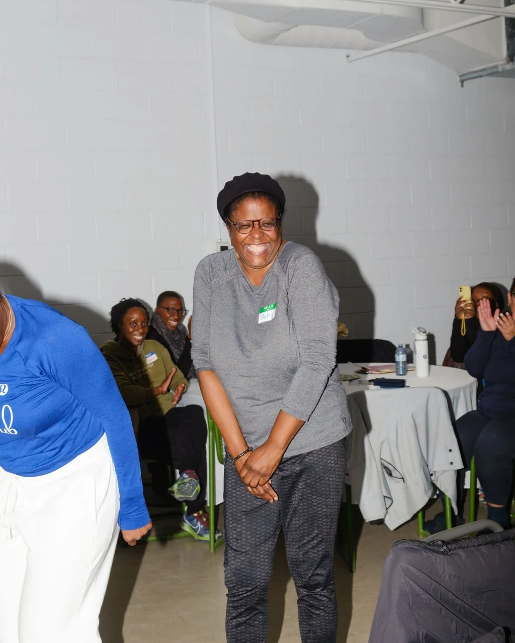 A woman smiling and laughing during a gathering indoors, with other women seated at tables in the background.
