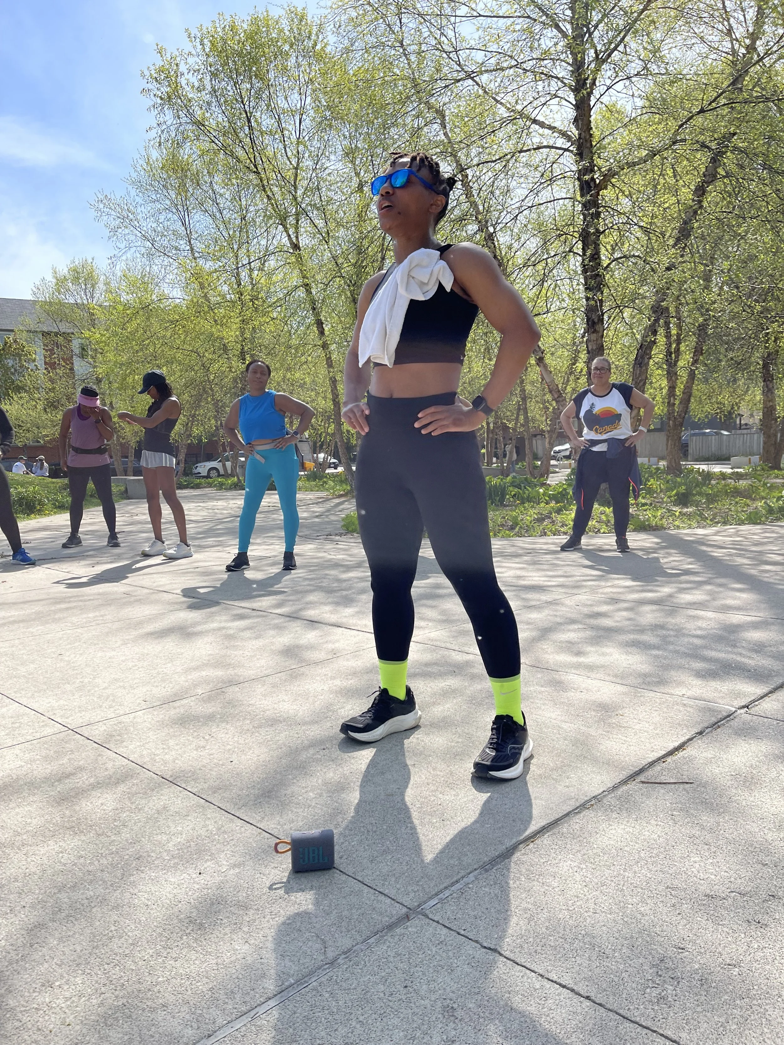 Group of women in workout clothes standing outdoors on a paved area with trees and buildings in the background, preparing for a fitness class or group exercise session. The woman in the foreground is wearing sunglasses, black athletic wear with yello