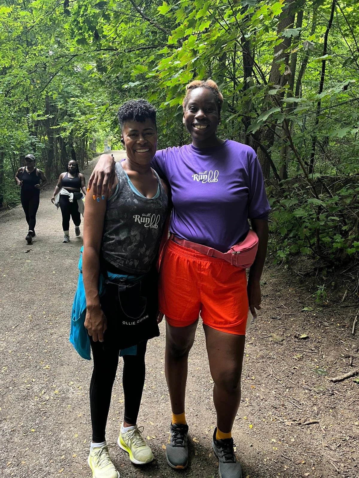 Two women smiling and standing together on a forest trail, with other people walking behind them. The women are dressed in sporty clothes, likely after a run or hike.