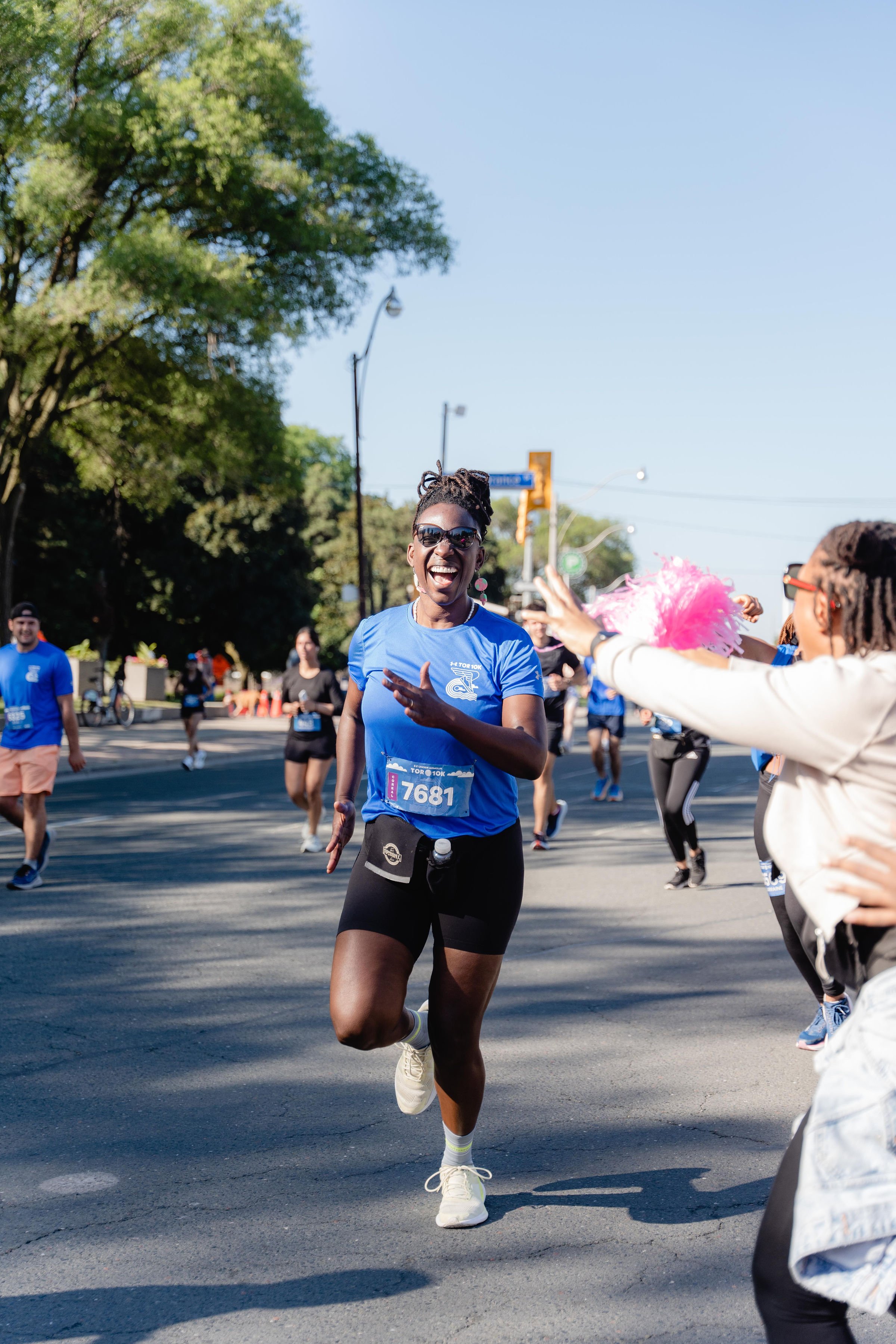 A woman wearing a blue race shirt, black shorts, and sunglasses smiling and running in a marathon while passing a woman with arms outstretched.