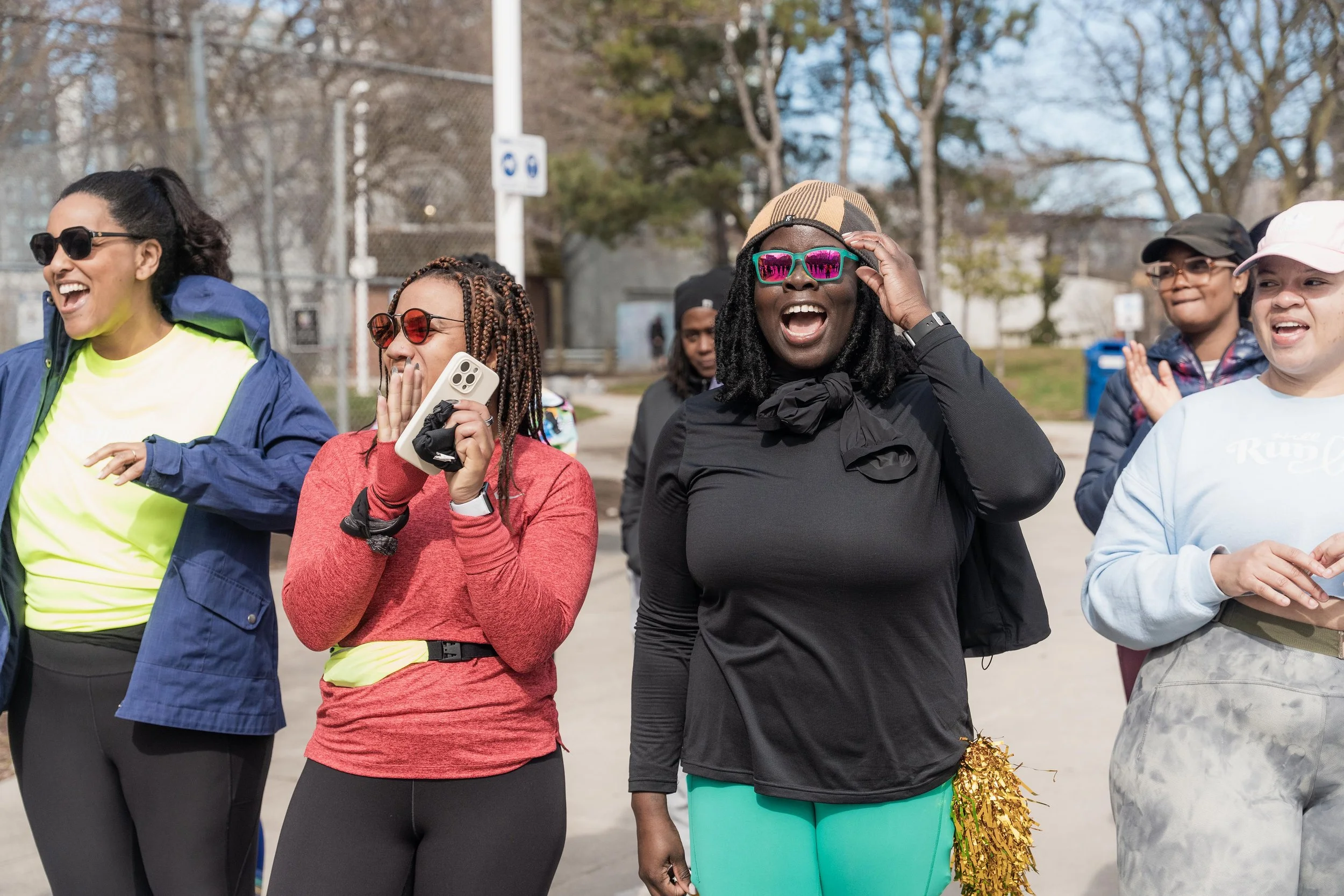 Group of women smiling and laughing outdoors, some wearing sunglasses and colorful athletic clothing, participating in a cheerful event or gathering during daytime.