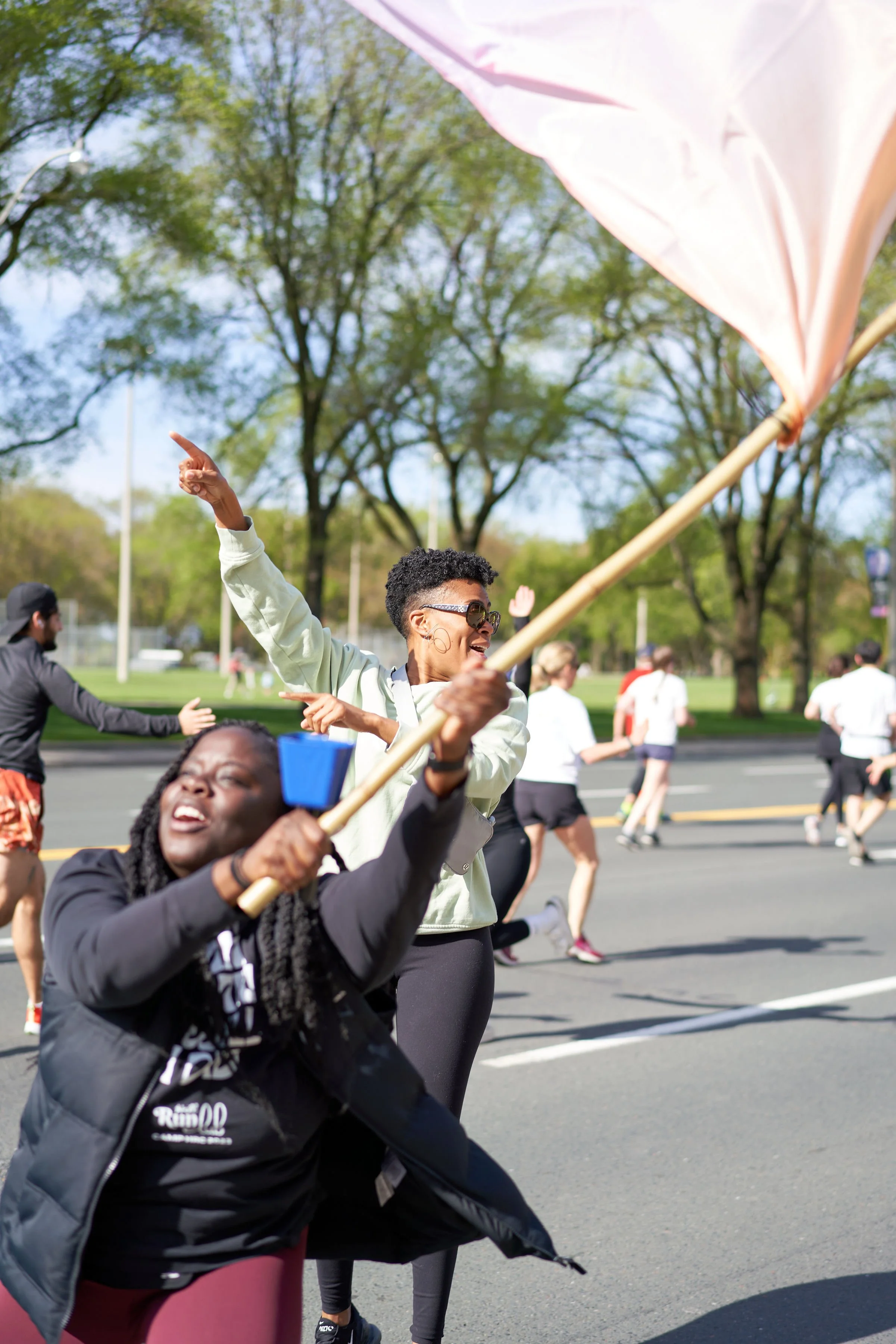People participating in a marathon race, with two women in the foreground holding flags and cheering, surrounded by runners on a tree-lined street on a sunny day.