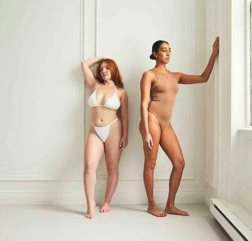 Two models wearing swimwear — one in a white triangle bikini, one in a nude one-piece bodysuit — standing against a white wall.