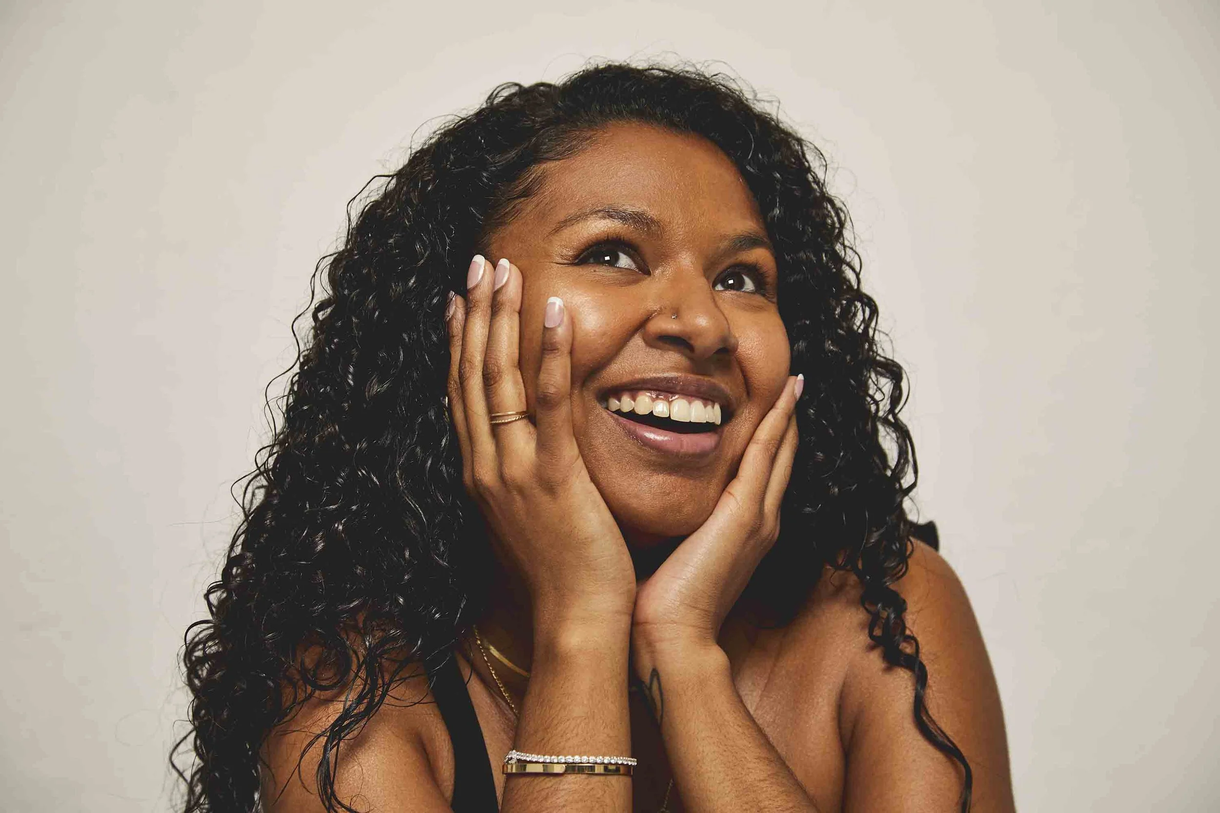 A woman with curly black hair smiling and touching her face with both hands against a plain white background.
