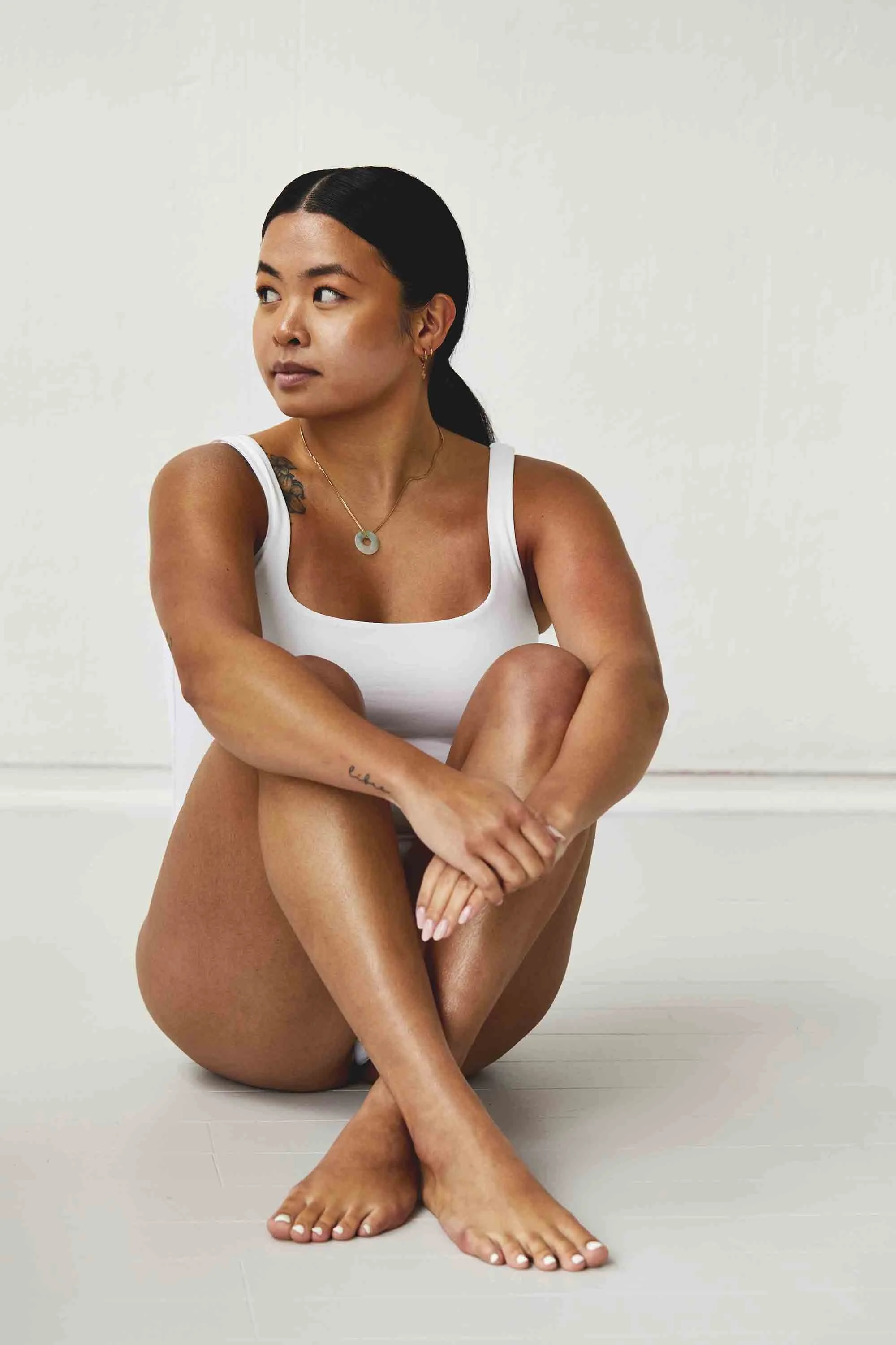 A woman sitting on the white floor, with her arms wrapped around her knees, looking to her left. She has black hair, tan skin, and is wearing a white tank top with a necklace and earrings. The background is plain white.
