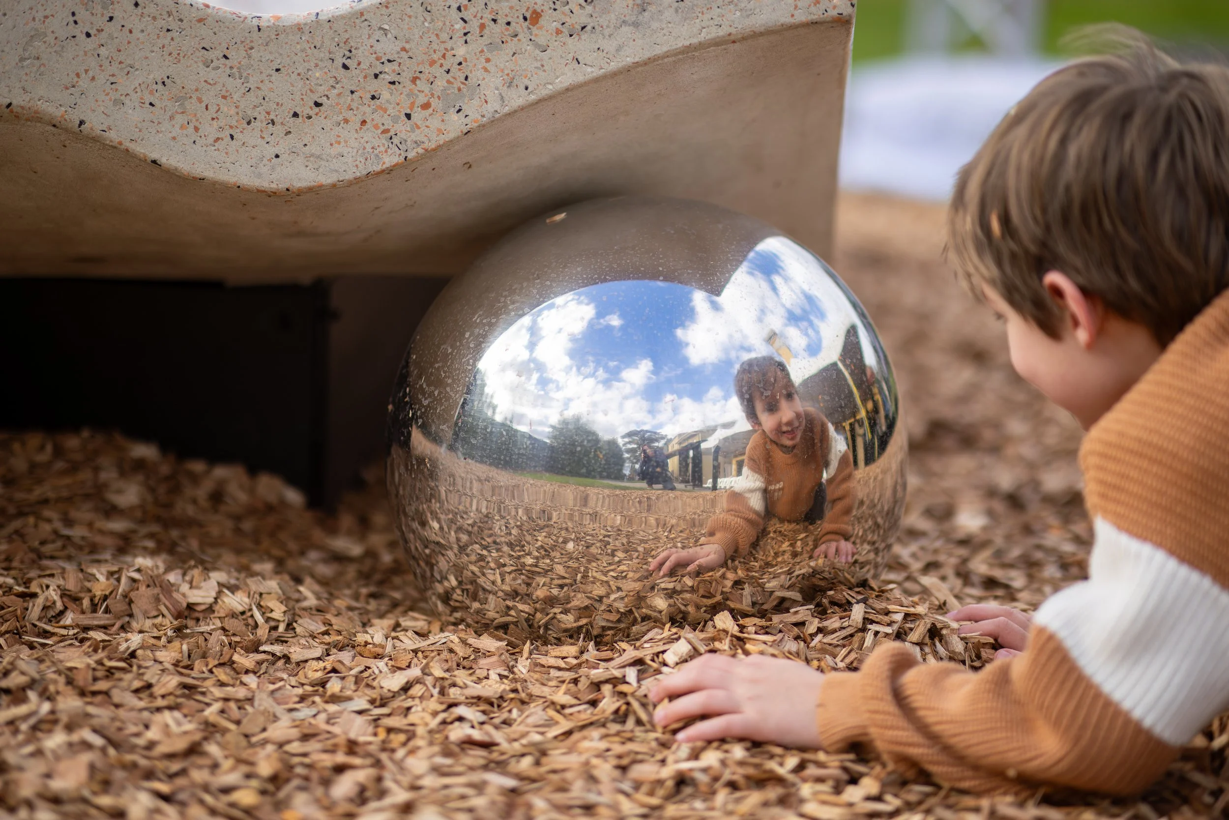 A young boy crouches on a bed of wood chips, looking at a shiny metal sphere that reflects the sky, clouds, and a person taking the photo.