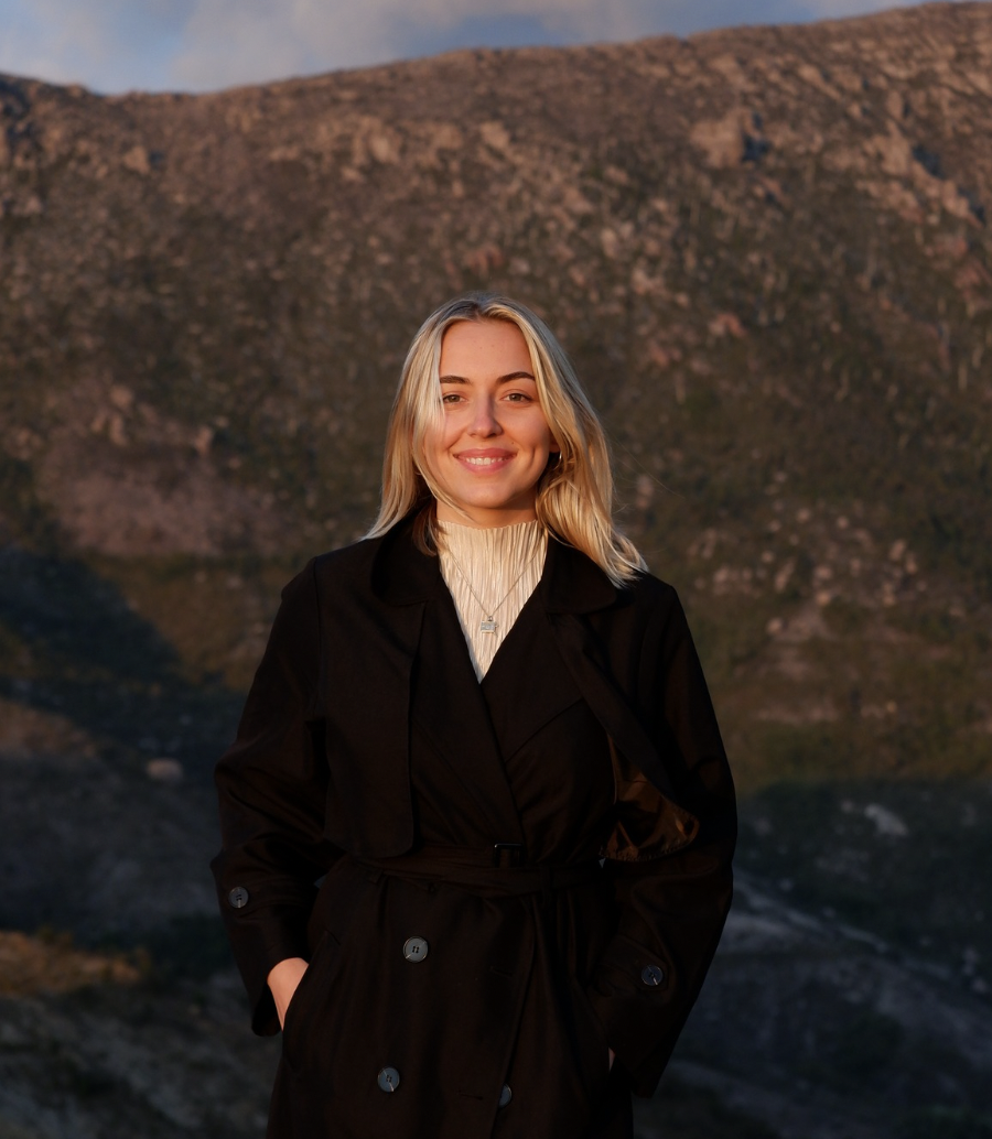 A smiling woman with blonde hair standing outdoors in front of a mountainous landscape during sunset.