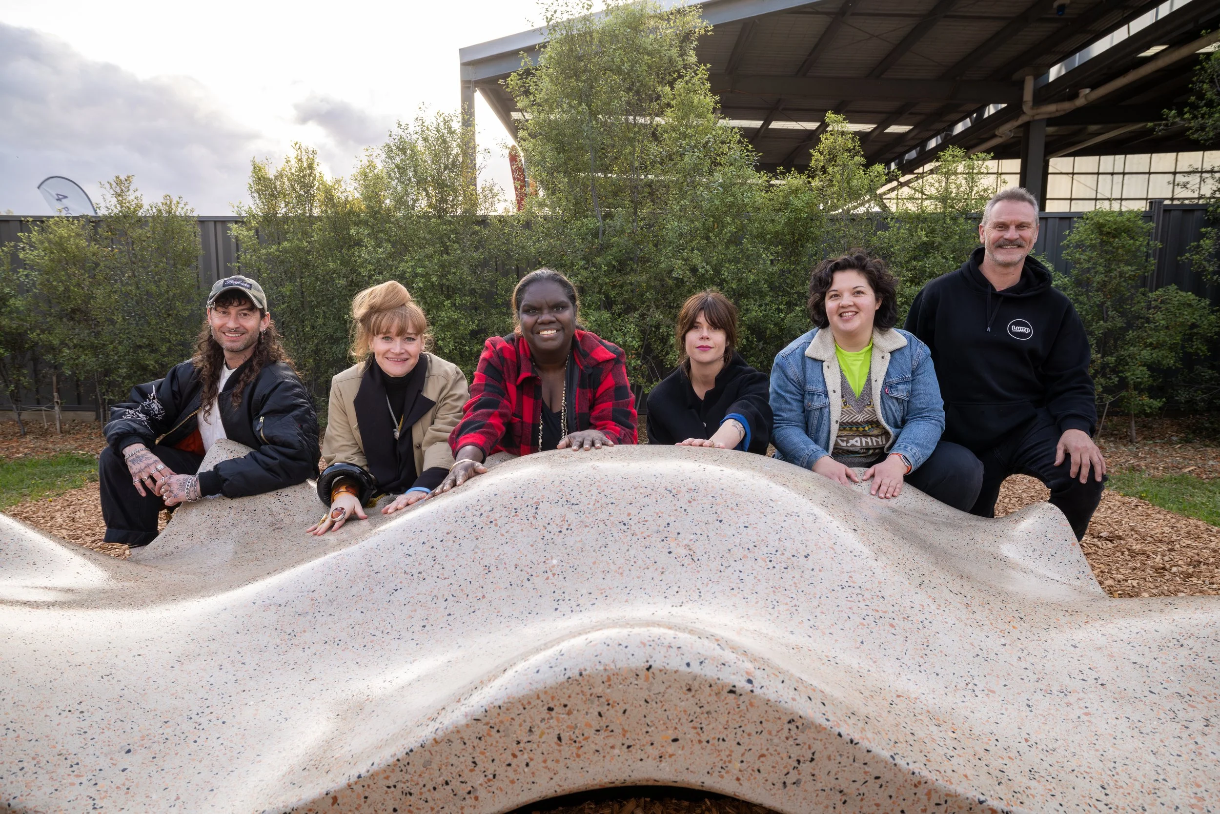 Group of six diverse people sitting on a curved, speckled concrete sculpture at an outdoor park, with trees and a large building with a metal roof in the background.