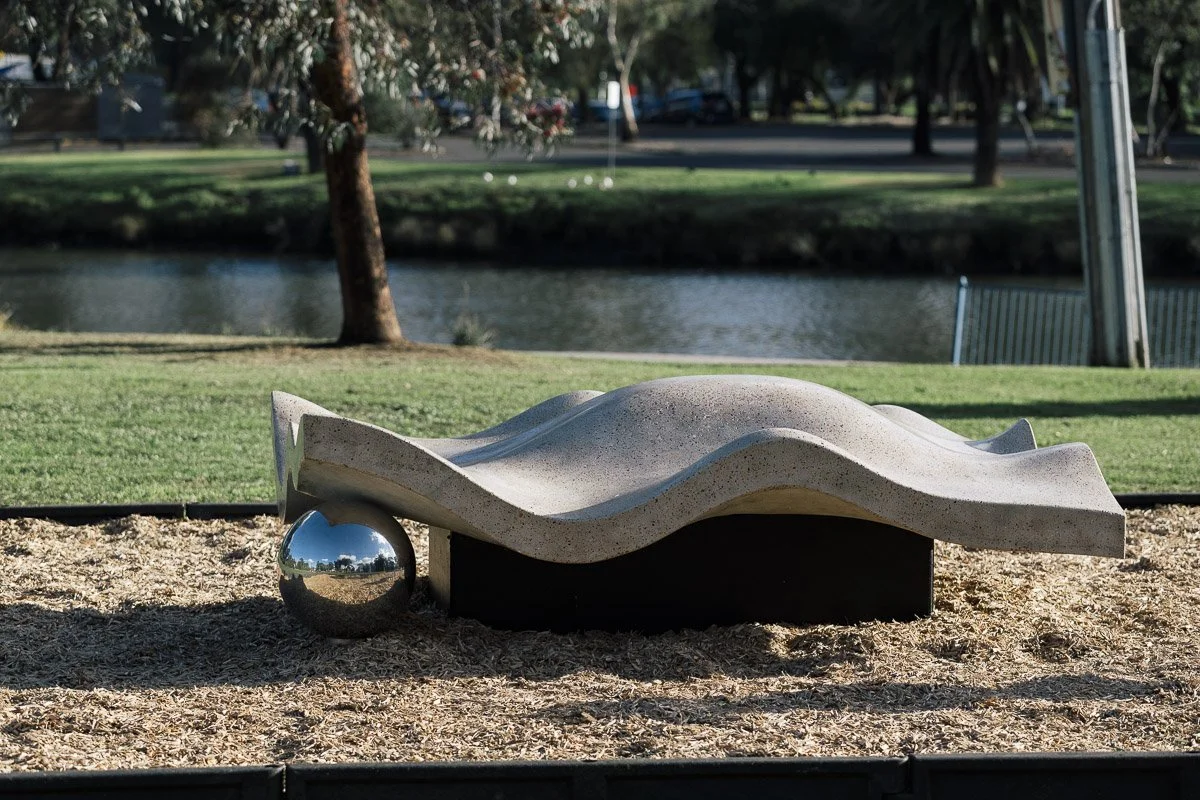 Modern sculpture of a reclining abstract figure with a shiny metal sphere at one end, situated outdoors on a patch of wood chips in a park near a pond with trees in the background.