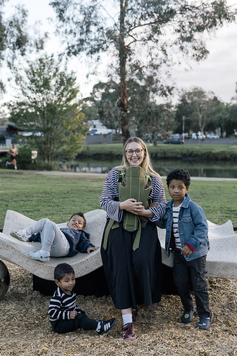 A woman with glasses holding a backpack, standing next to two young boys, one sitting on a bench and the other standing, with a third boy sitting on the ground, in a park with trees and a pond in the background.