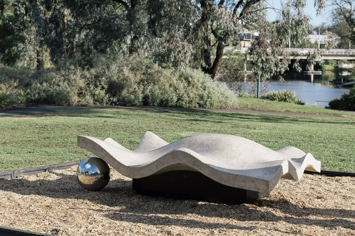 Abstract modern outdoor concrete bench with a reflective metallic sphere at one end, situated on a sandy surface in a park with greenery, trees, and a river in the background.