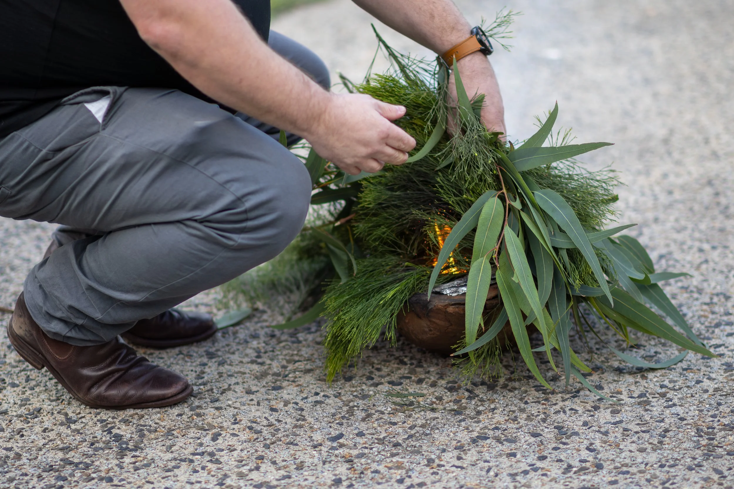 A person kneeling on a gravel surface, arranging a green foliage and branches in a large flowerpot.