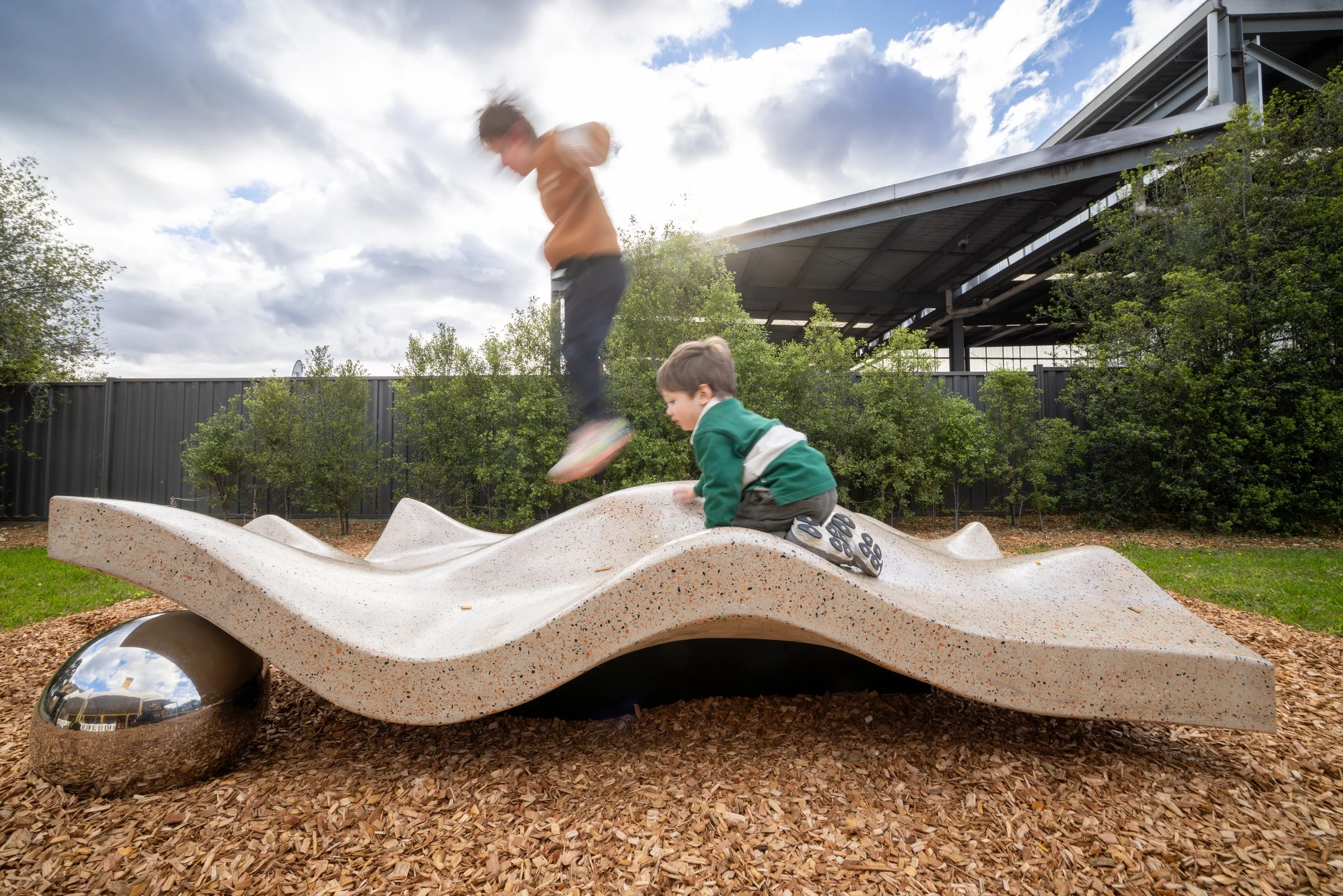 Two children playing on a modern playground structure with a textured surface, located outdoors under a cloudy sky.