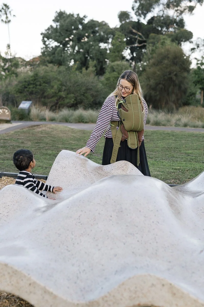 A woman and a young child at a park, with the woman smiling and touching a sculpted, wave-like structure, while the child looks on. The woman is wearing glasses, a striped shirt, and a backpack, and the child is in a striped shirt. There are trees an