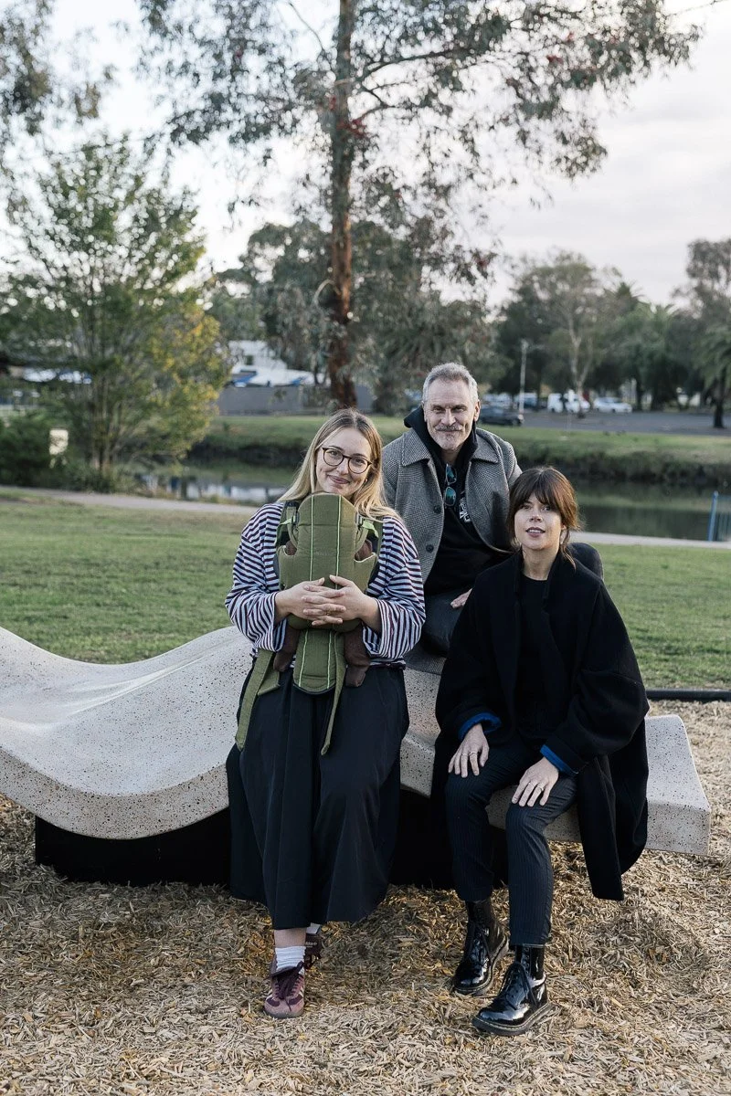 Three people in a park with trees, a pond, and a cloudy sky. One woman is sitting on a bench, another woman standing with a backpack, and a man standing behind them.