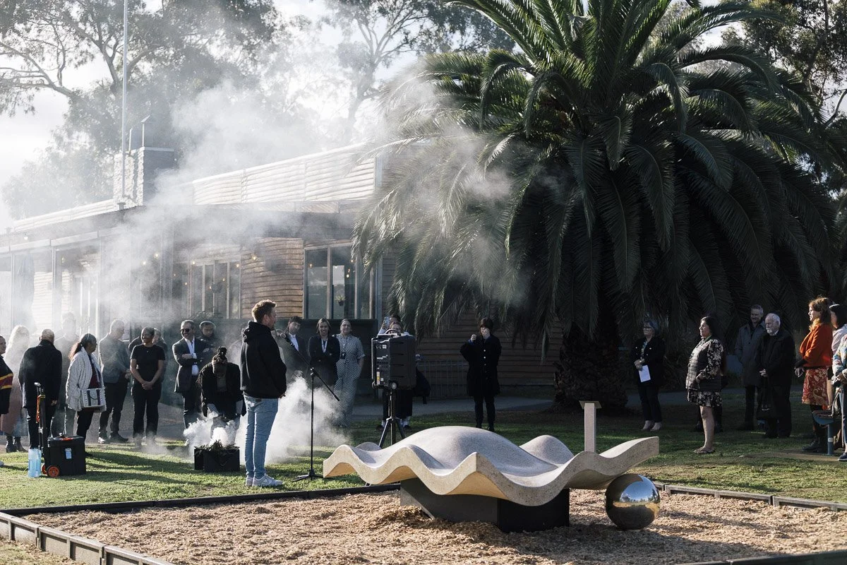 A person speaking into a microphone to a crowd gathered outdoors near a large palm tree and a modern building, with smoke or fog effects, during daytime.