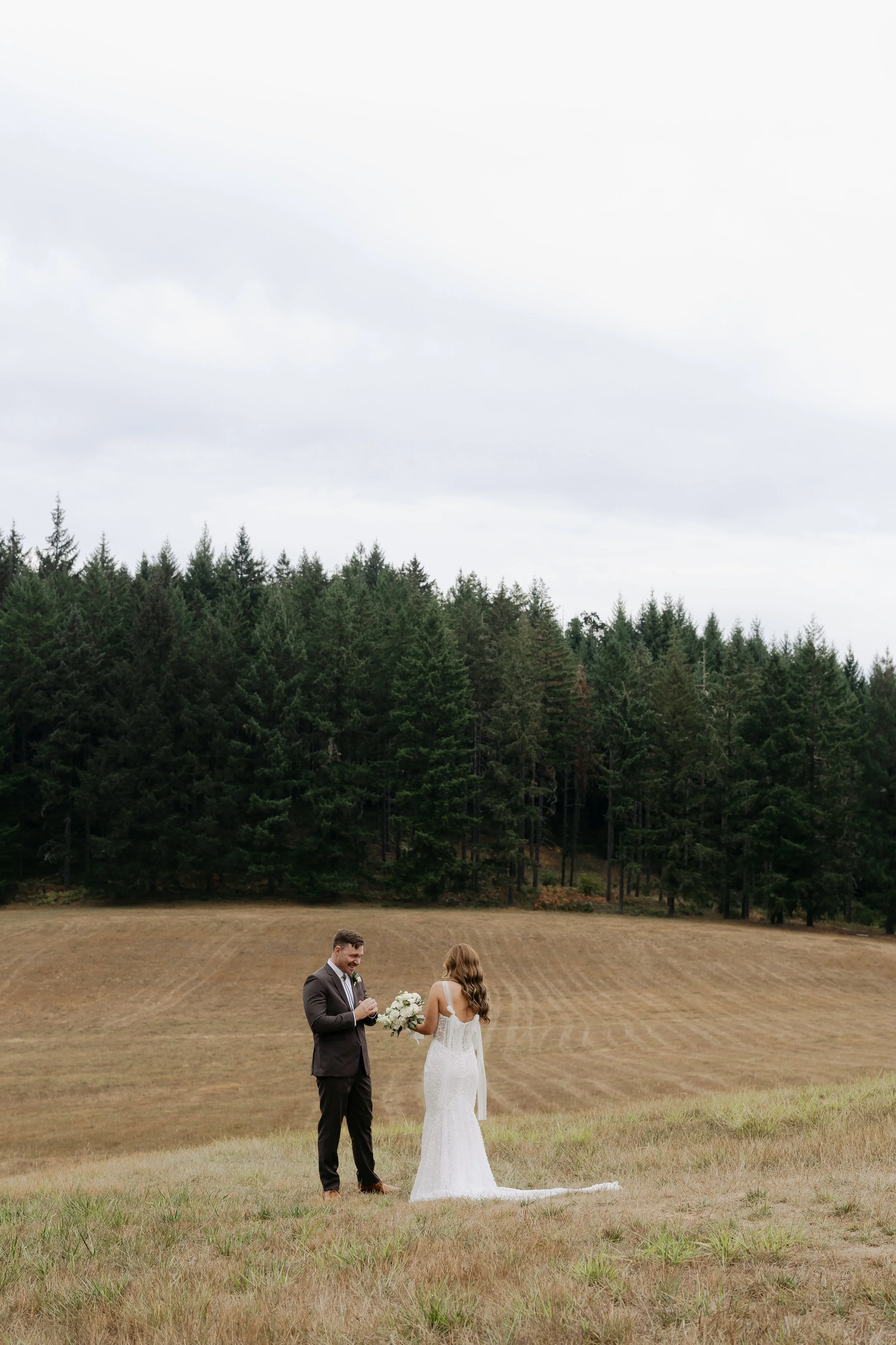 A bride and groom stand on a field with a forested hillside in the background. The groom is adjusting his jacket while the bride holds a bouquet, and they are facing each other in a wedding ceremony outdoors on an overcast day.