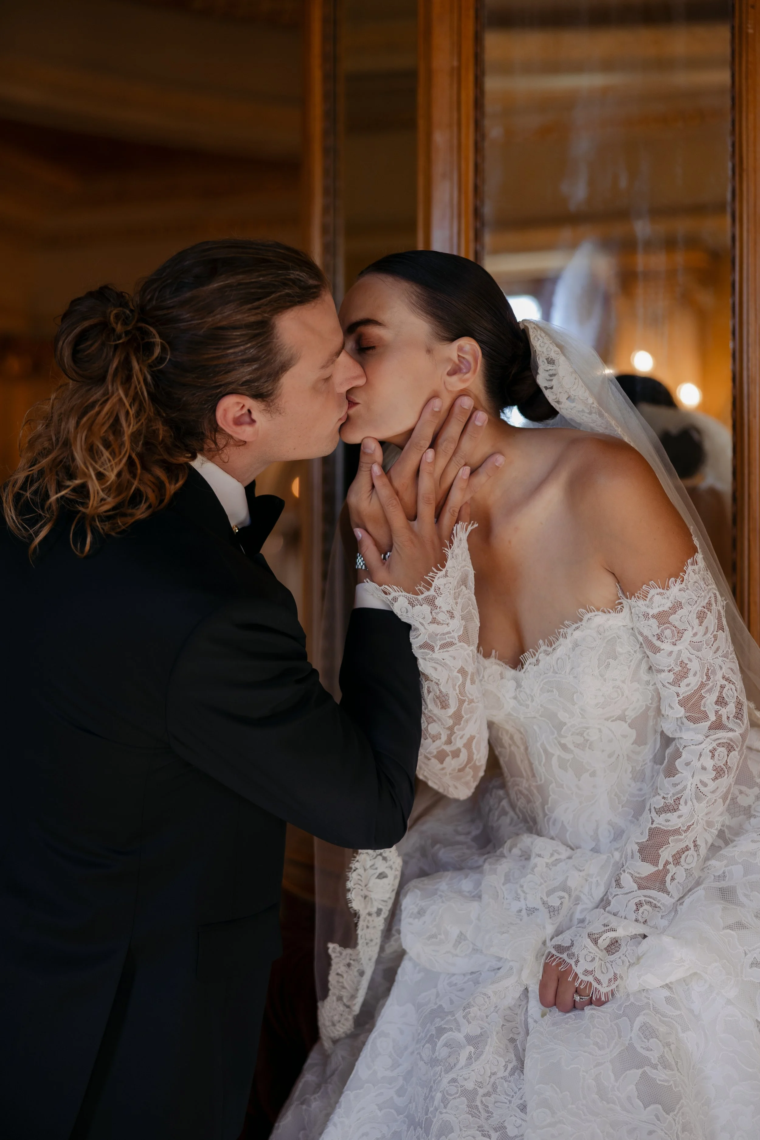 A bride and groom are sharing a kiss in front of a mirror. The groom has long, wavy hair in a bun and is wearing a black tuxedo. The bride has dark hair styled in a sleek updo, is wearing a white lace wedding dress with long sleeves and a veil, and i