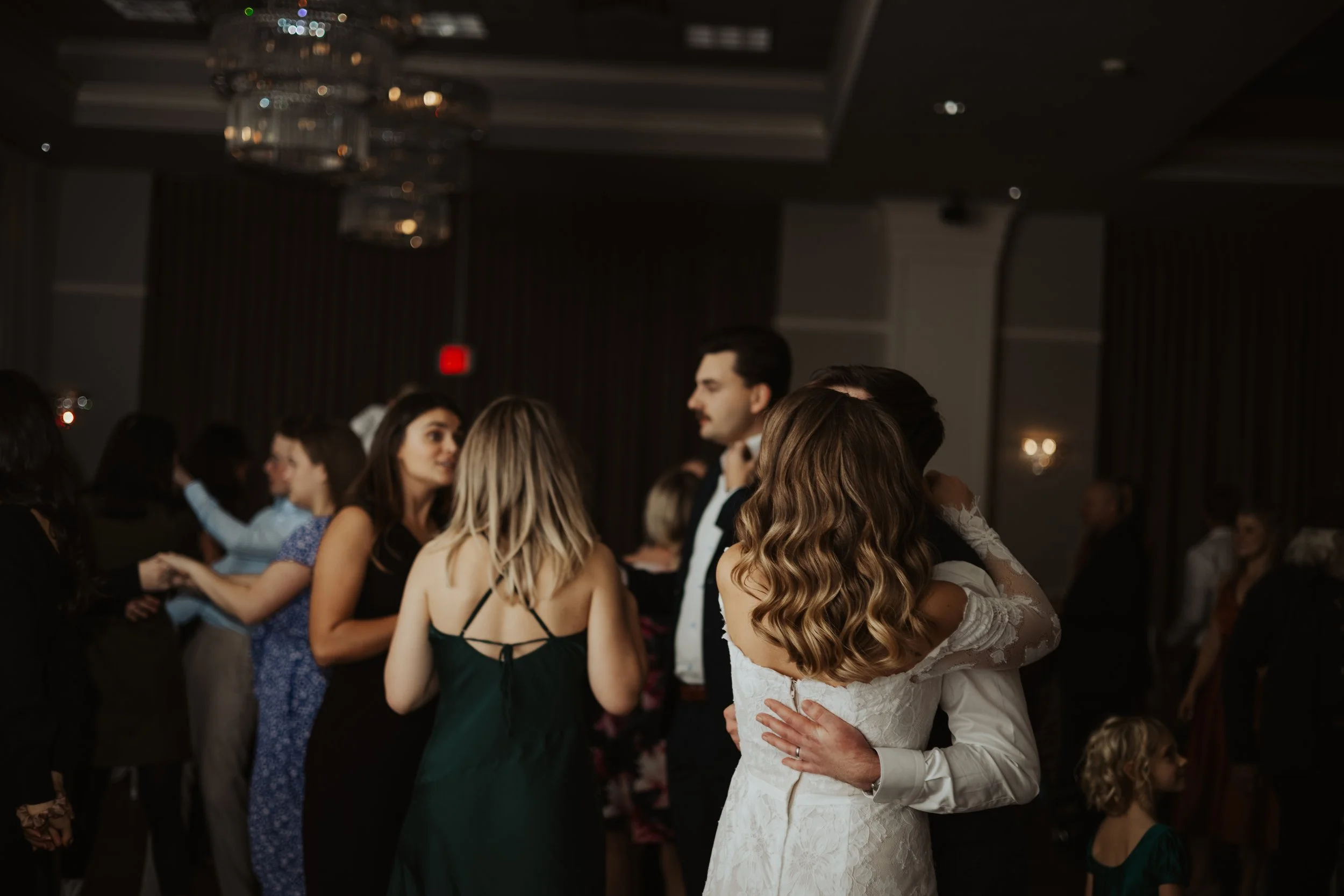 A bride and groom are dancing closely at a wedding reception, surrounded by other guests on the dance floor, in a dimly lit elegant ballroom with chandeliers.