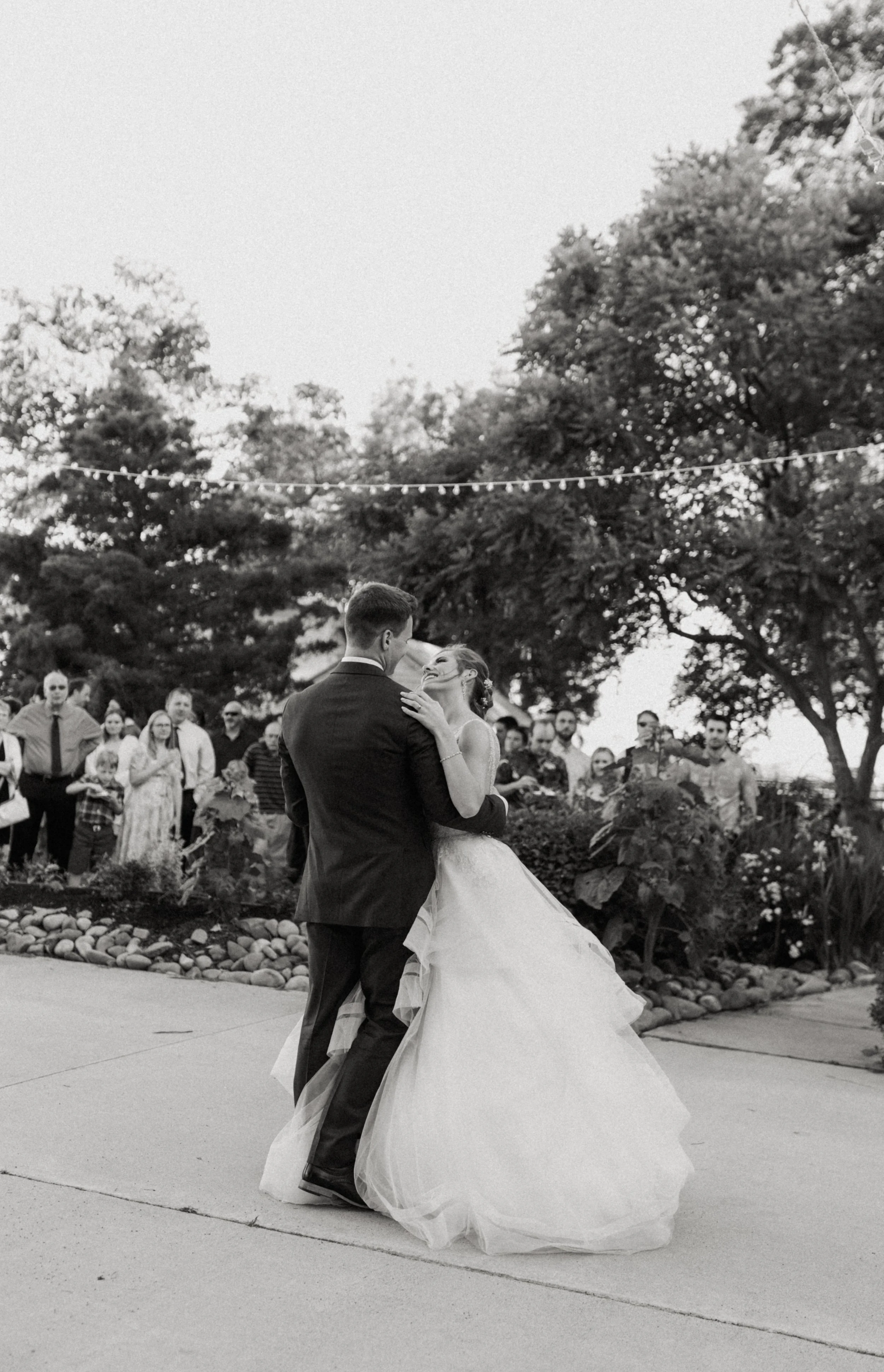 Couple dances outdoors at wedding reception, surrounded by guests and trees, black and white photograph.