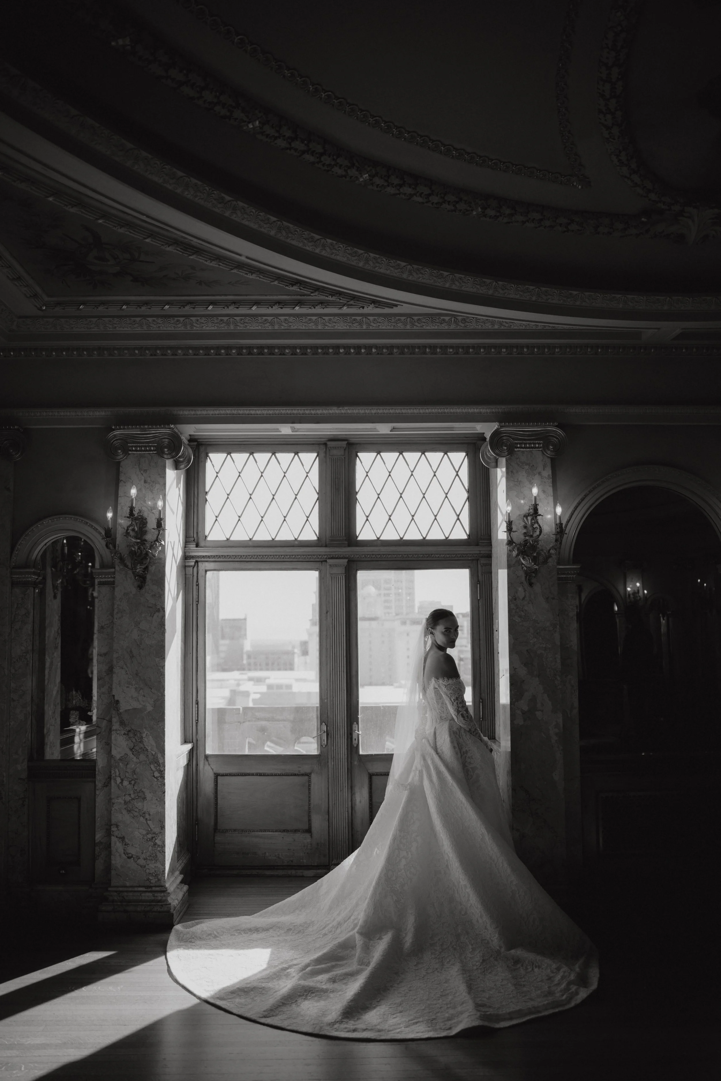 A bride in a wedding gown standing near a large window, with sunlight creating shadows on the floor, in an ornate room with decorative ceiling and wall details.