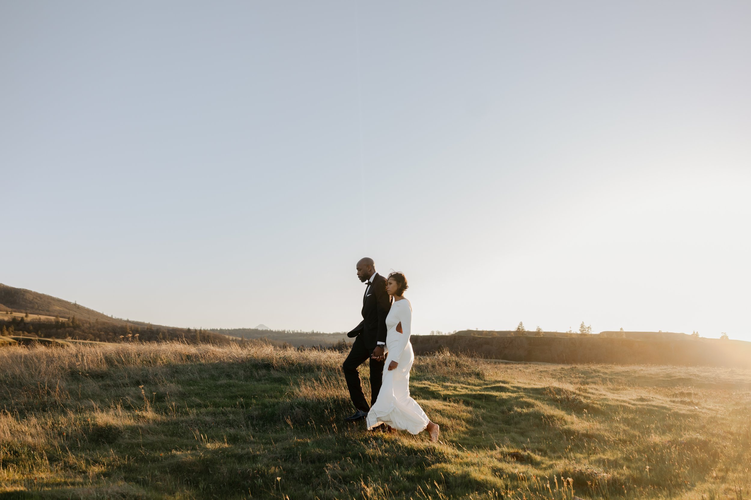 A man and woman walking hand in hand across a grassy field during sunset, with rolling hills in the background.