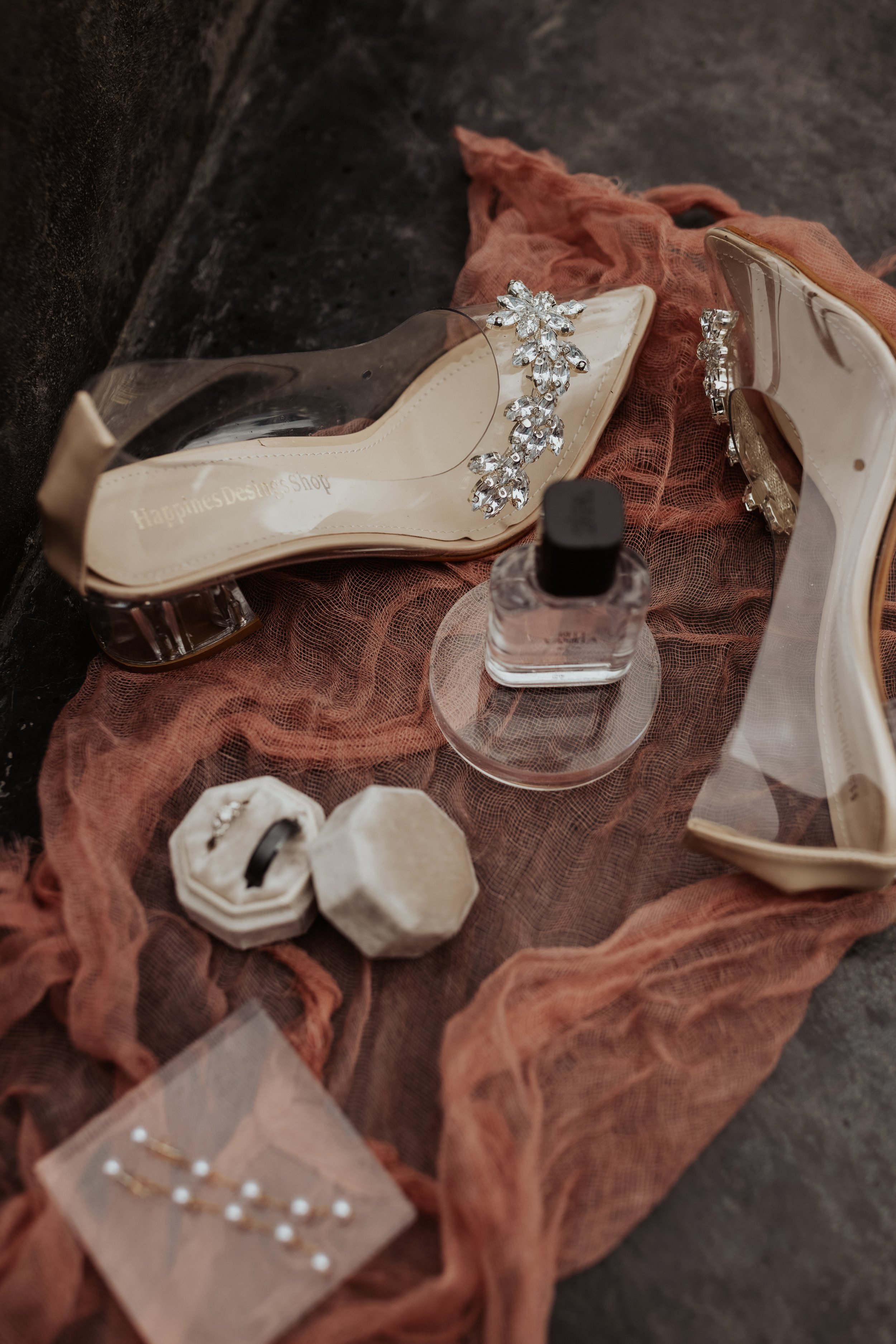 Elegant bridal accessories including rhinestone shoes, pearl and diamond jewelry, perfume bottle, and rings on soft pink fabric.