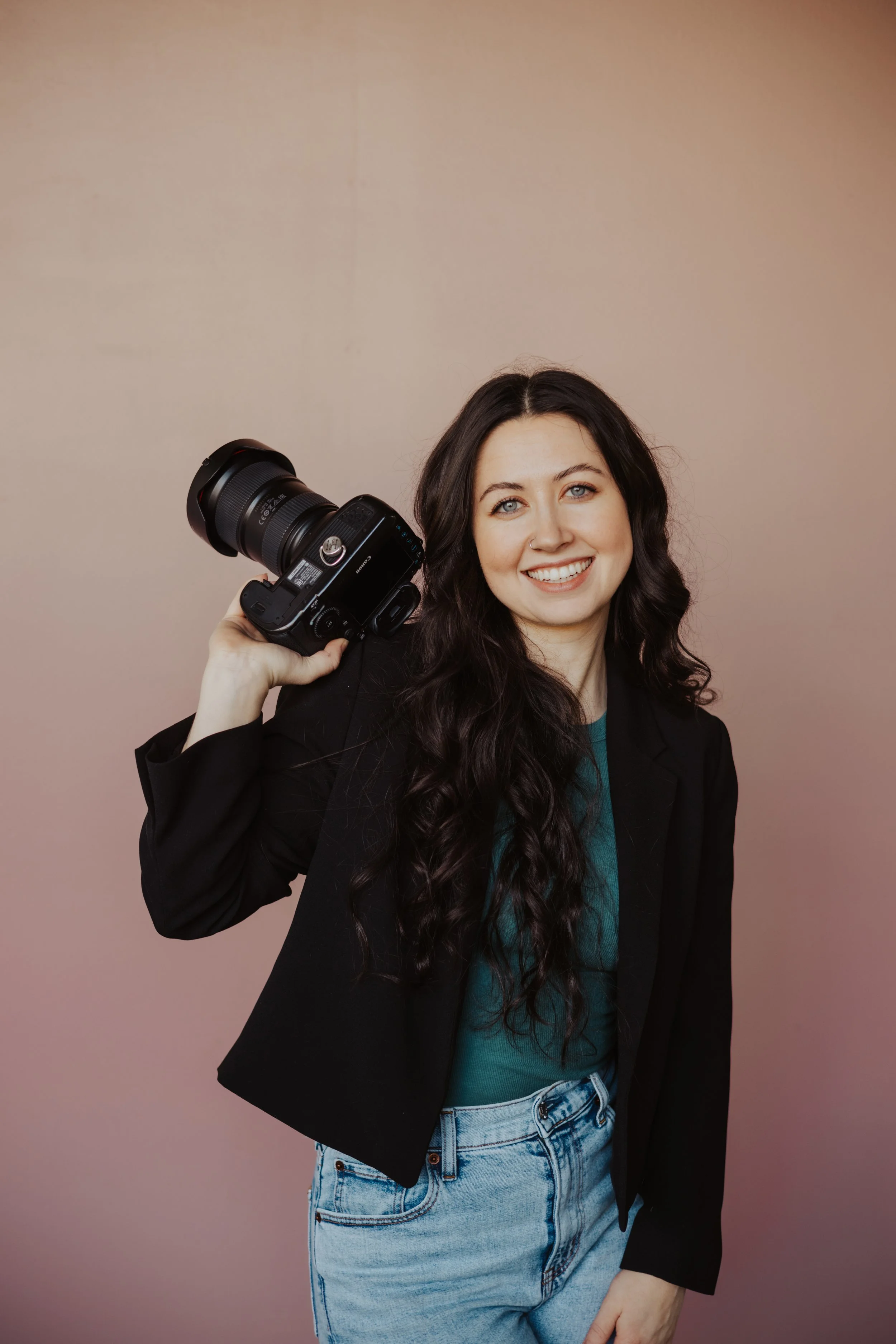 A young woman with long wavy dark hair and blue eyes holding a professional camera with a large lens, smiling, wearing a black blazer over a teal top and high-waisted light blue jeans, standing against a plain beige background.