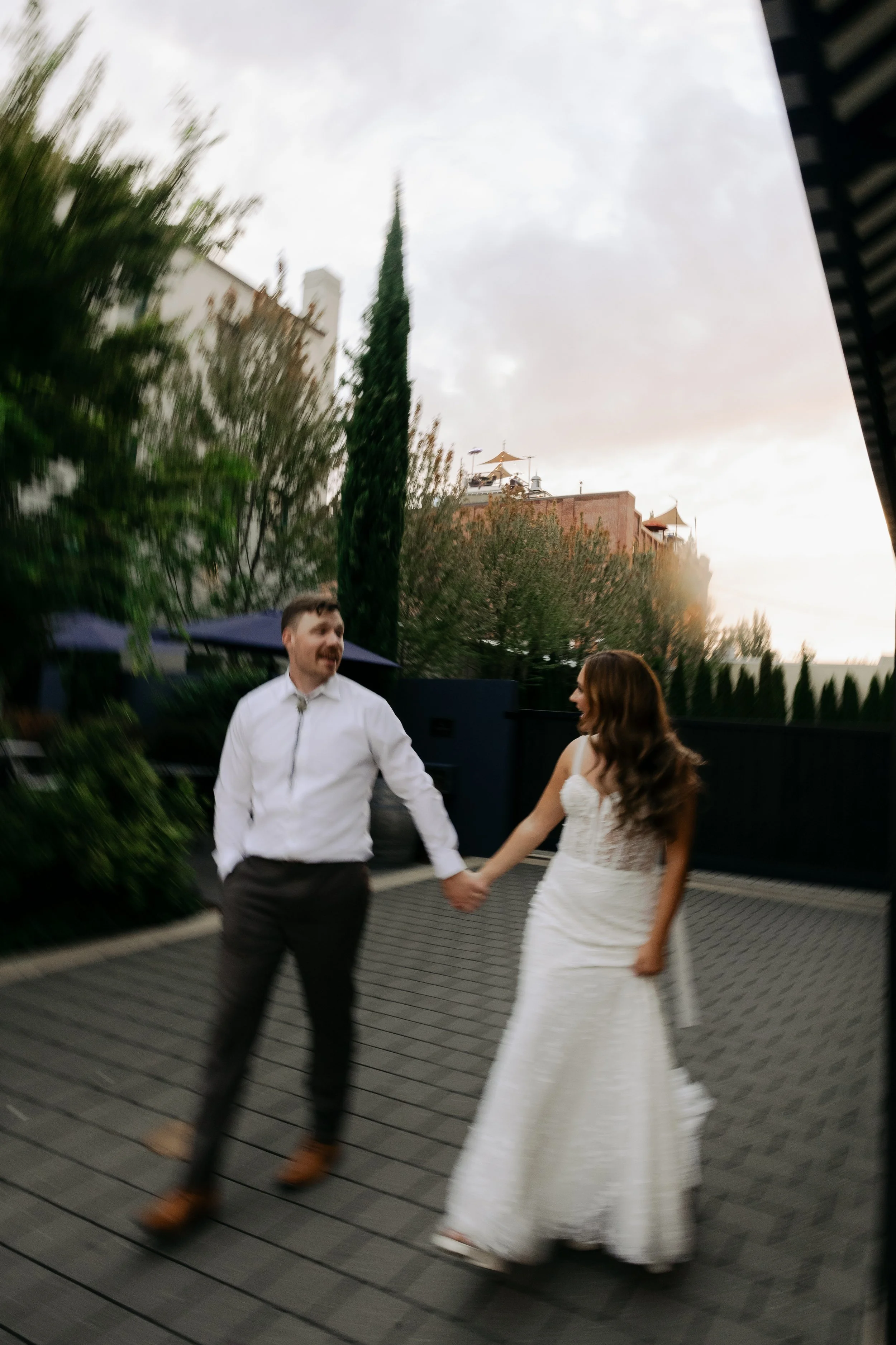 A man and woman holding hands, dressed for a wedding, walking on a tiled outdoor area. The man is wearing a white shirt and dark pants, while the woman is wearing a white strapless wedding gown.