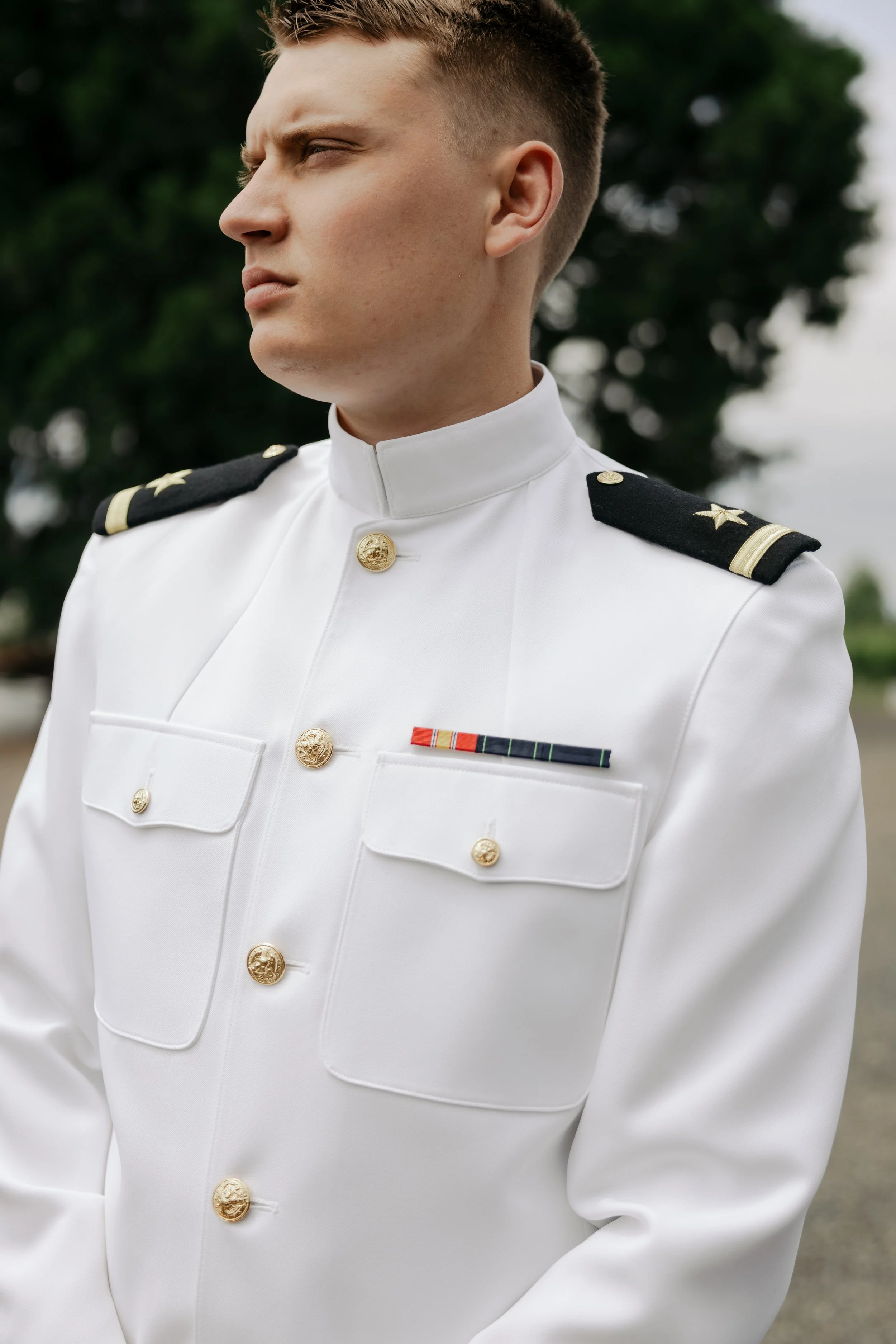 Close-up of a young man in a white military dress uniform with medals, standing outdoors with blurred trees in the background.