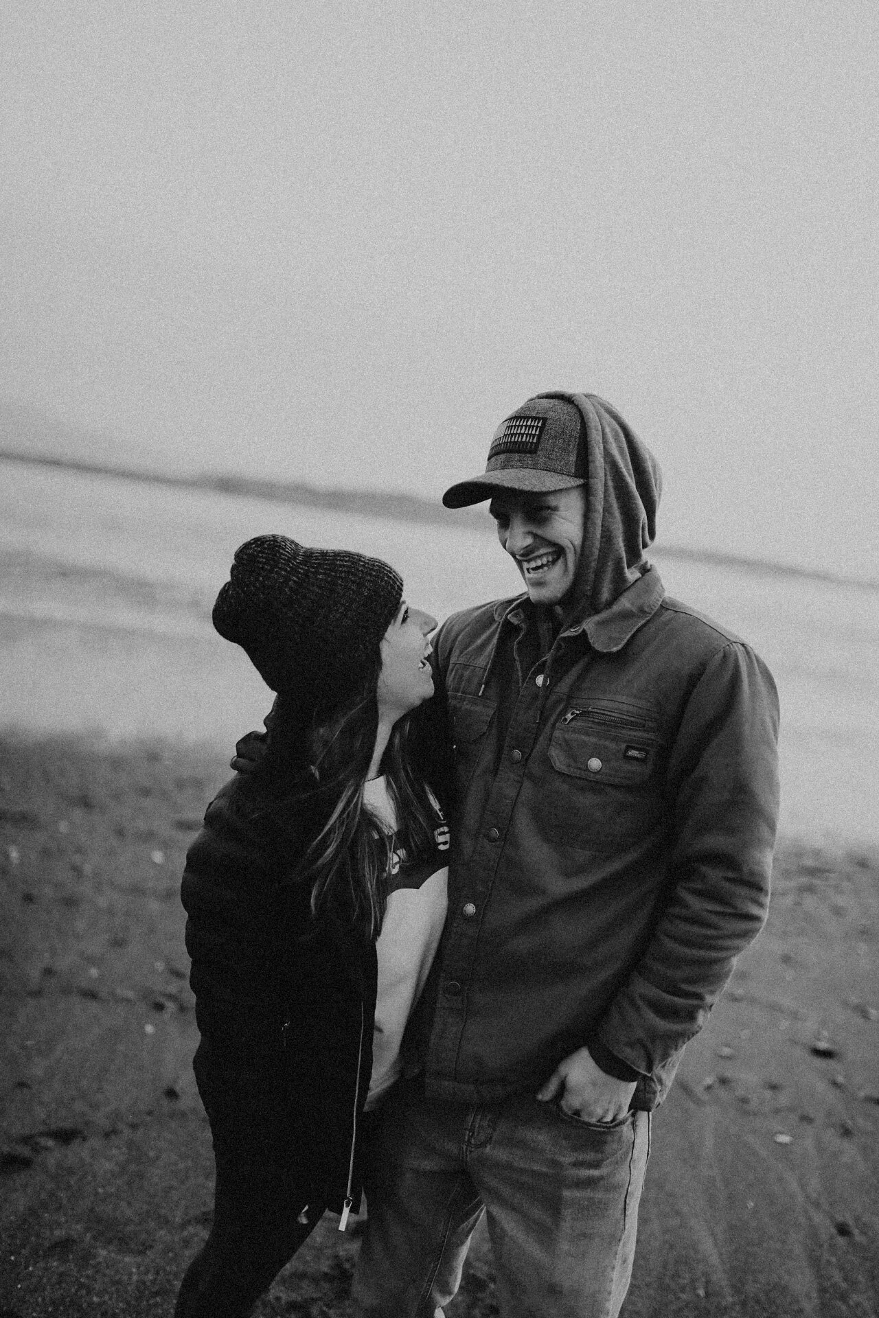 A black and white photo of a smiling couple on a beach, looking at each other affectionately. The woman wears a beanie and hoodie, and the man wears a cap with a hoodie underneath, a jacket, and jeans.