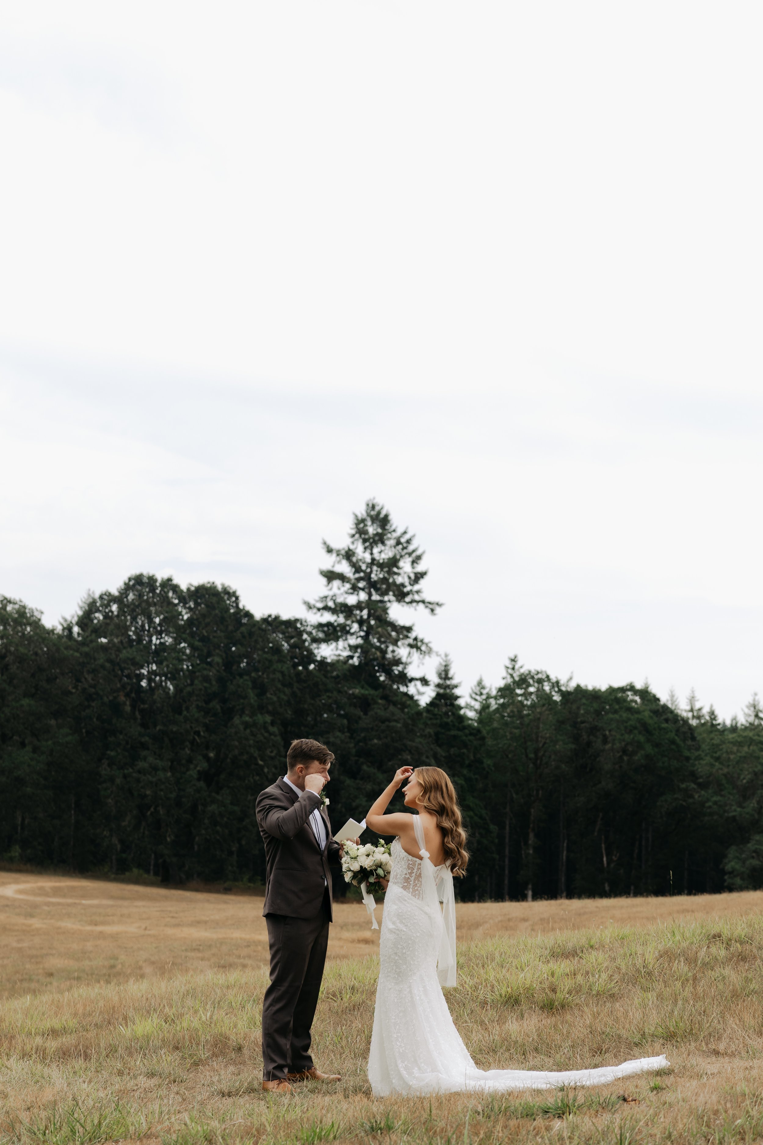 Bride and groom standing in an open field, facing each other, with the bride holding a bouquet of flowers. The groom appears emotional, with one hand near his face, while the bride is smiling and touching her hair. A forested area is in the background under a cloudy sky.