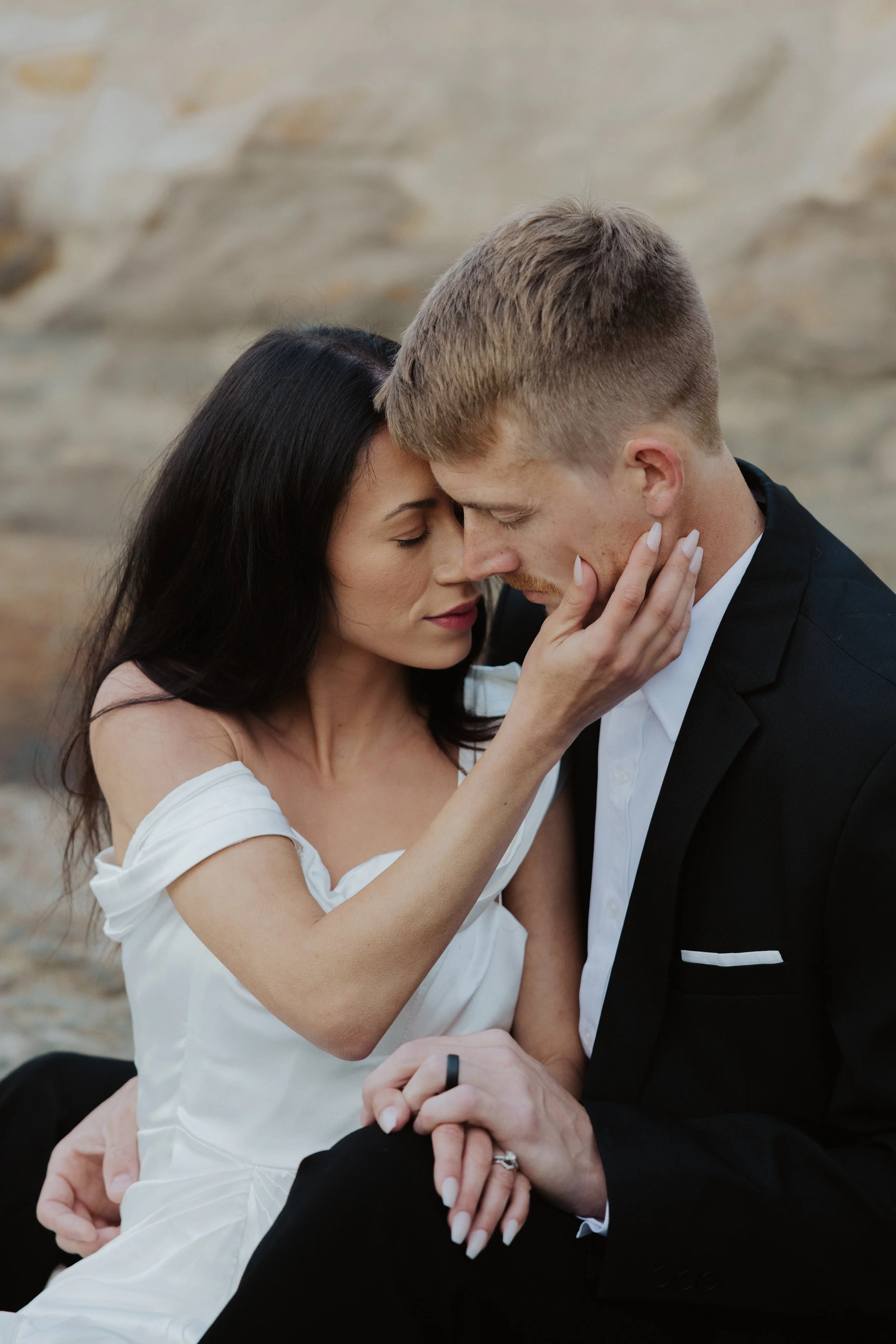 A romantic moment between a couple, with their foreheads touching and eyes closed, outdoors with a rocky background. The woman is gently holding the man's face and has dark hair, while the man has short, light hair and is wearing a black tuxedo.