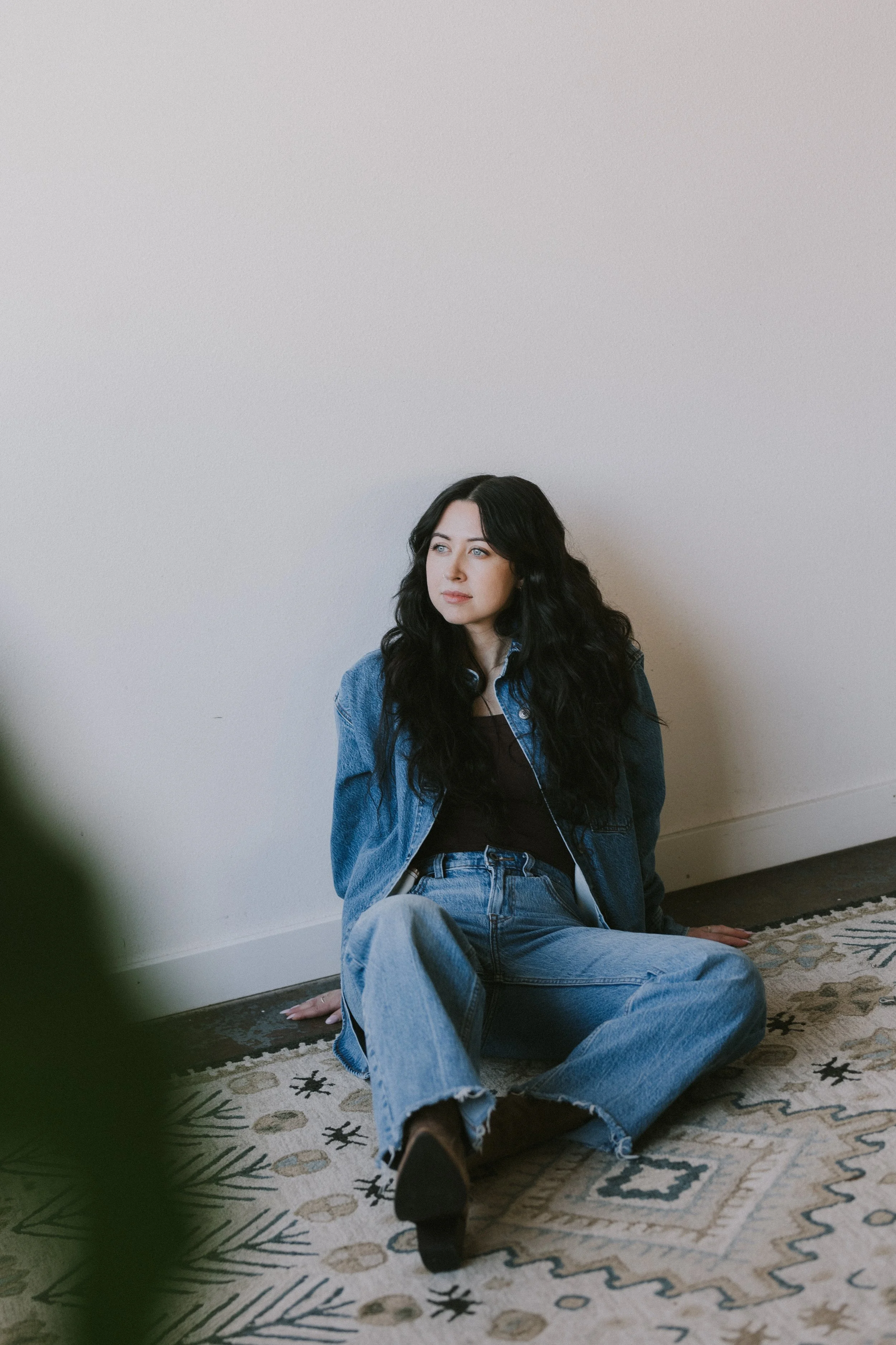 A woman with long black wavy hair sitting on the floor against a plain white wall, wearing a denim jacket and jeans, looking to the side.