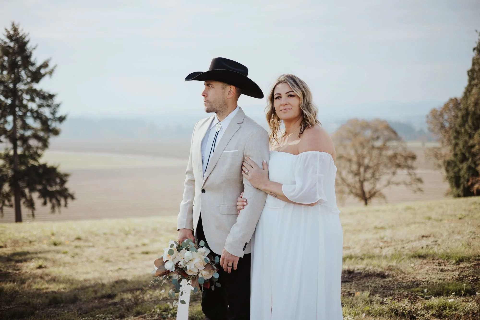 A bride and groom standing outdoors in a rural landscape, with the bride holding onto the groom's arm, both looking thoughtfully into the distance. The groom is wearing a light-colored blazer, a white shirt, a bolo tie, and a black cowboy hat. The br