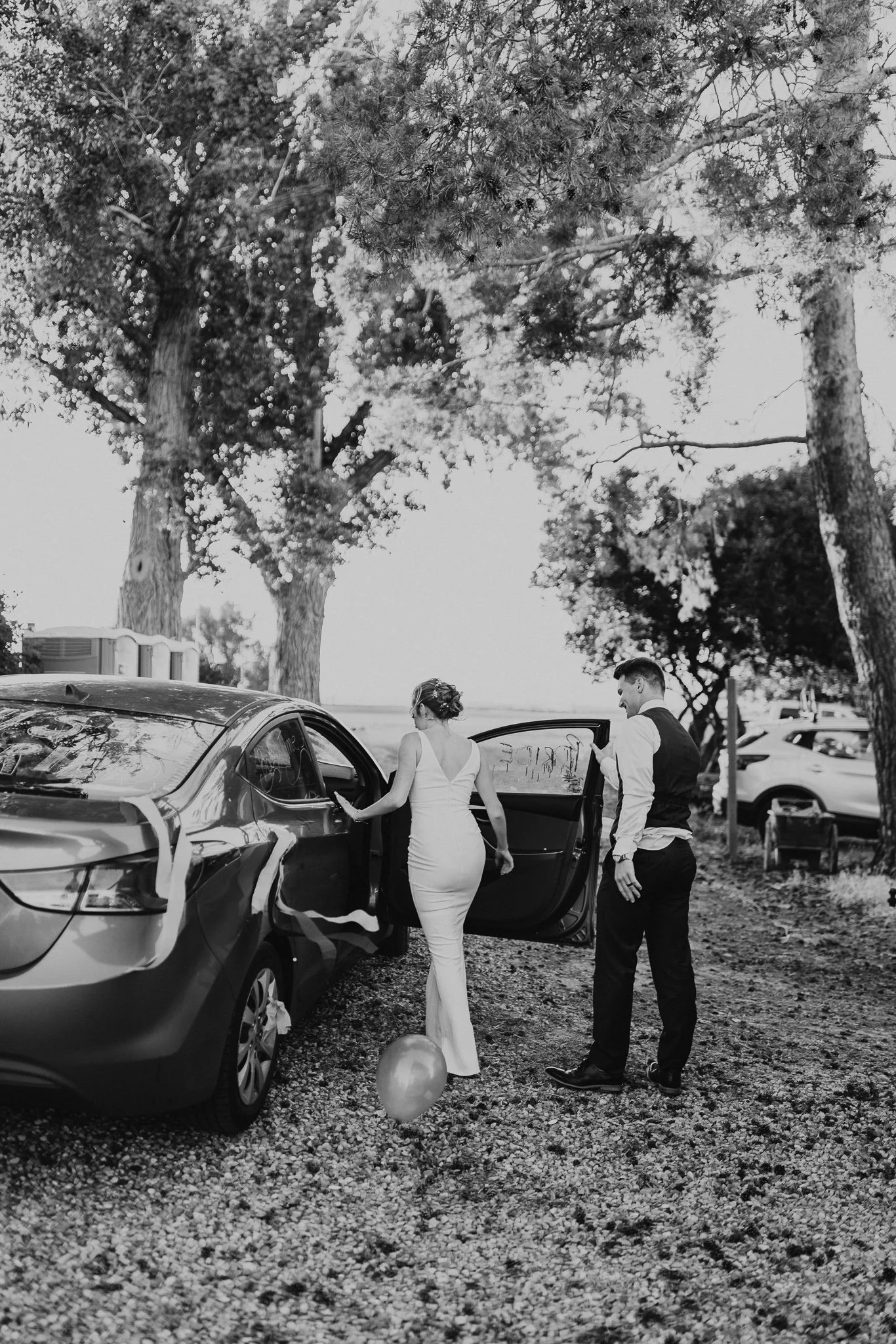 A woman in a white dress and a man in a vest and dress pants standing by a car, outdoors with trees and parked cars in the background.