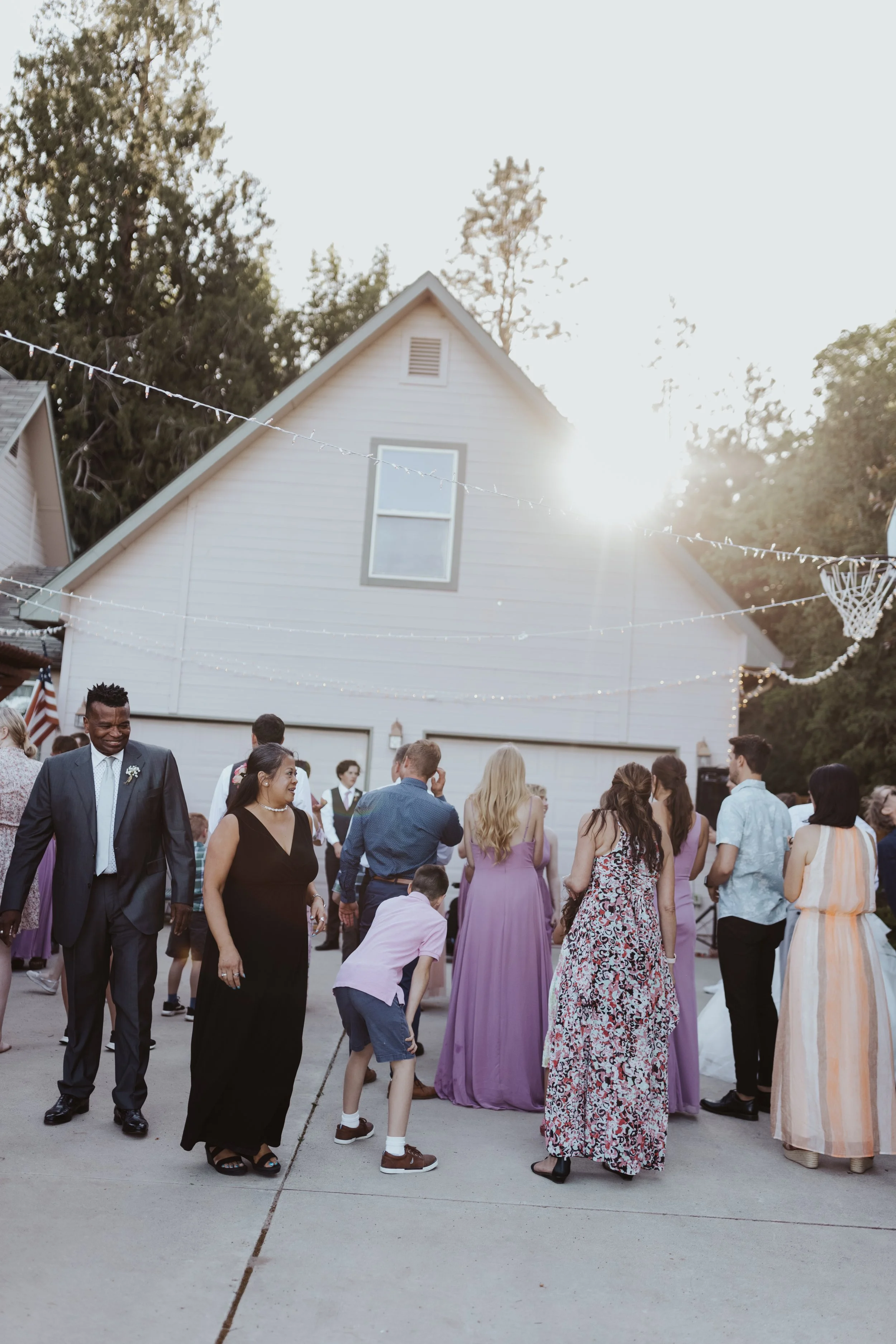 People gathered outside in front of a white house decorated for an outdoor celebration, with string lights in the sky and the sun shining brightly.