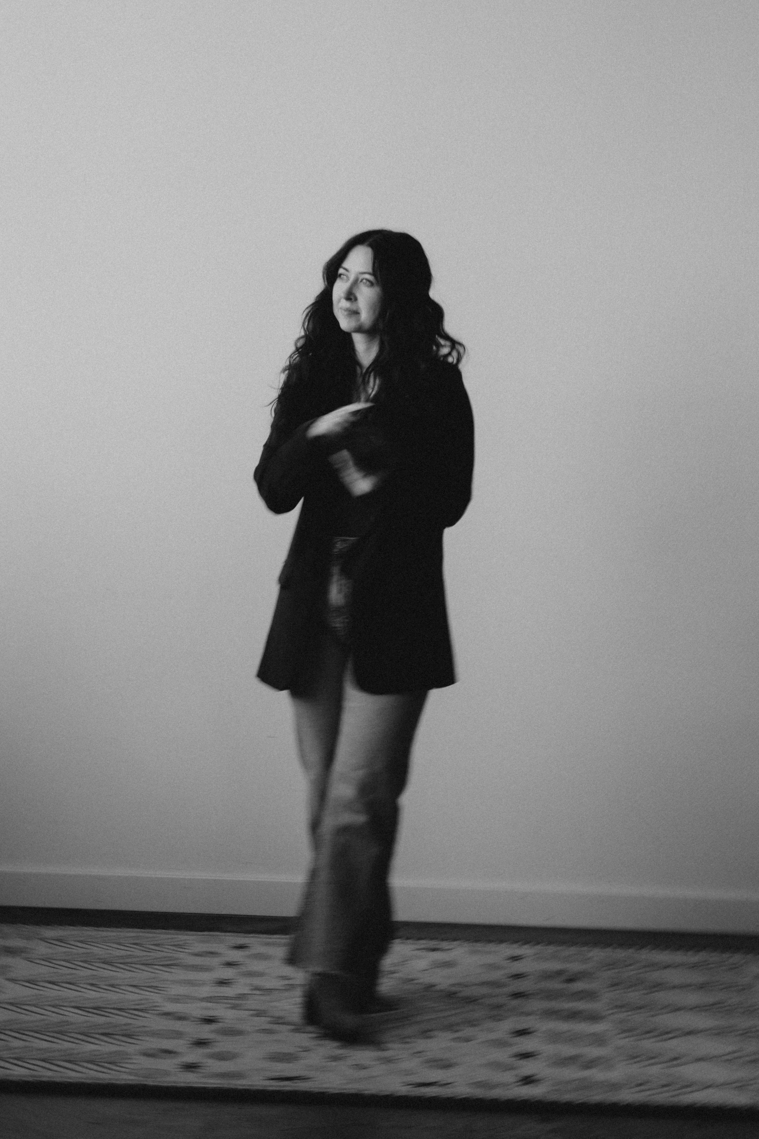 A woman with long, wavy dark hair standing on a patterned rug against a plain wall, dressed in a dark blazer, shiny black top, flared jeans, and boots, looking thoughtfully to the side.