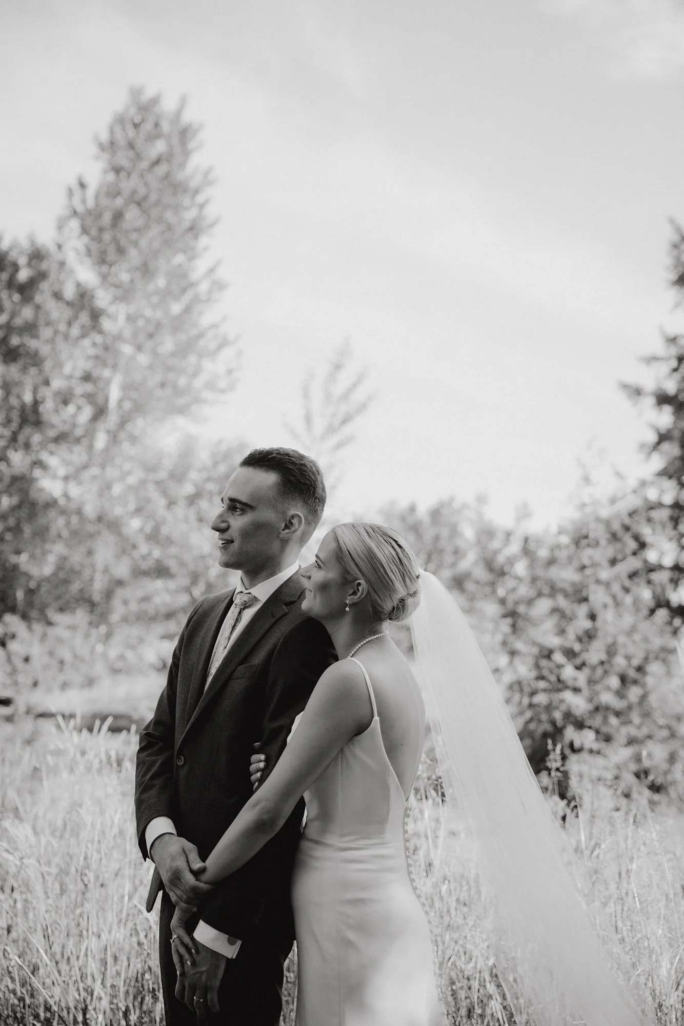 A black and white photo of a bride and groom standing outdoors, the bride leaning on the groom's shoulder with her eyes closed. The groom is smiling slightly and looking away. The bride wears a sleeveless wedding dress with a veil, and the groom wear