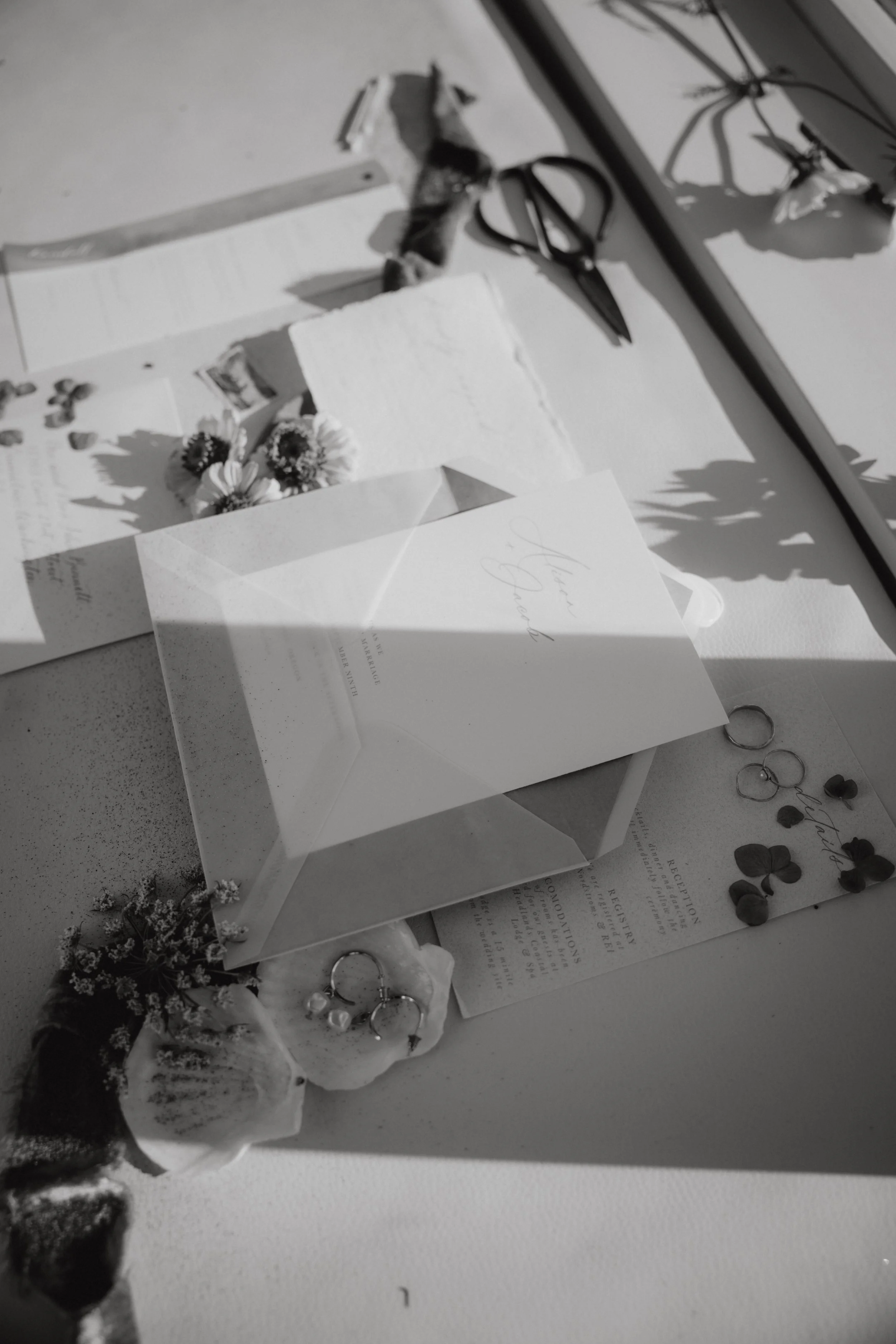 A wedding flat lay with wedding invitation, rings, flowers, and scissors on a white surface, with sunlight creating shadows.