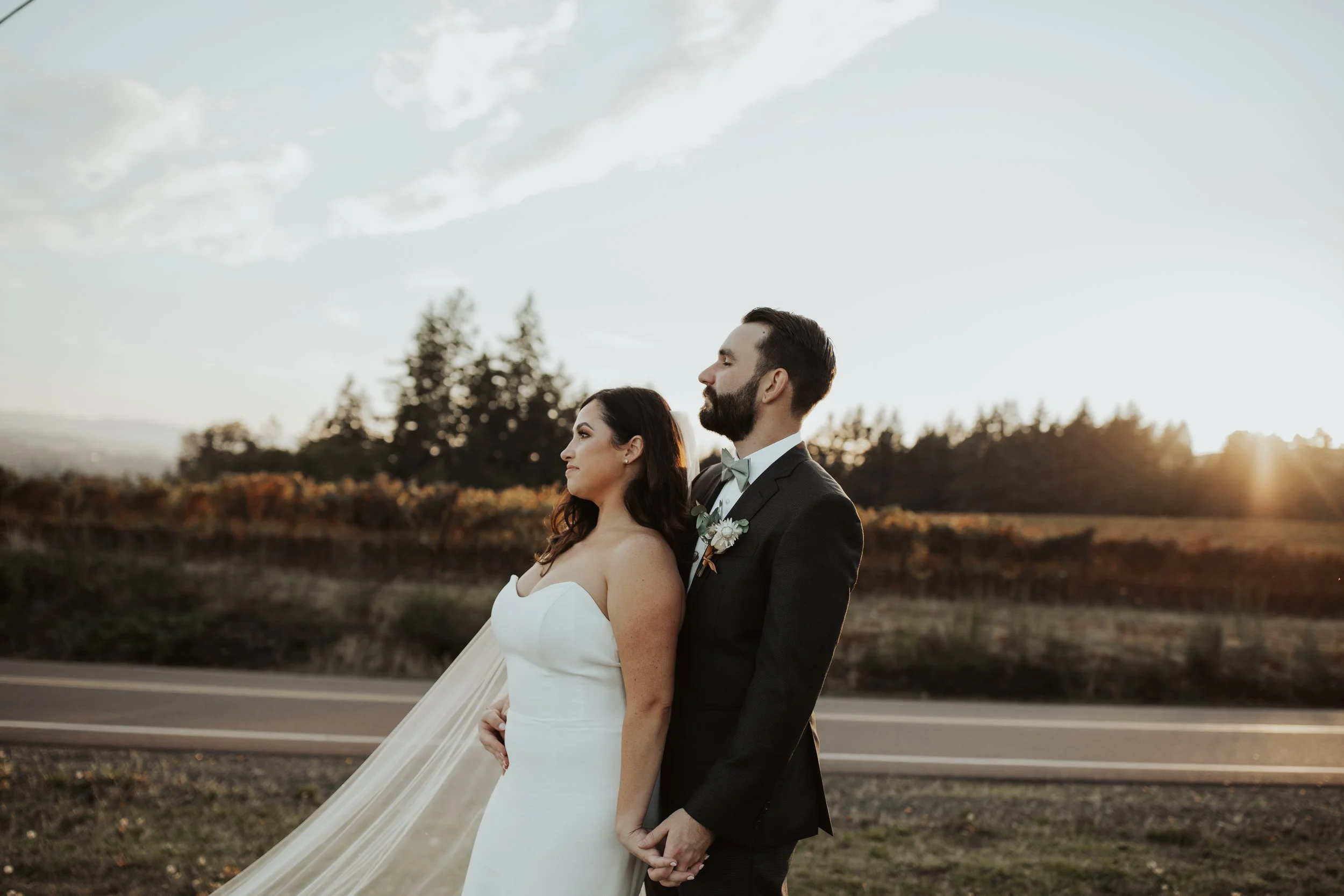 A bride and groom standing outdoors at sunset, holding hands, with a scenic landscape in the background. 