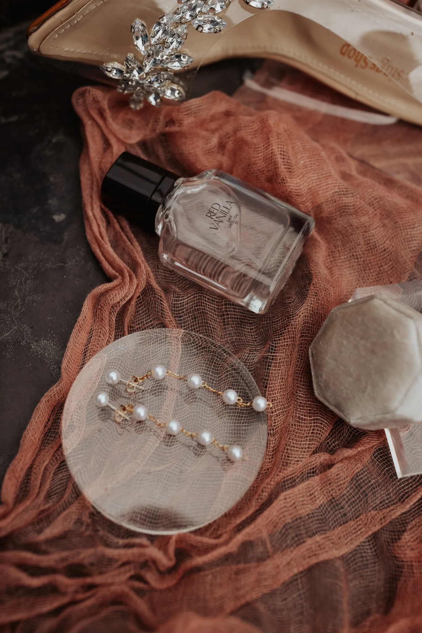 A bottle of perfume, pearl earrings on a transparent dish, a velvet jewelry box, and a sheer pink fabric with a sparkly shoe in the background.