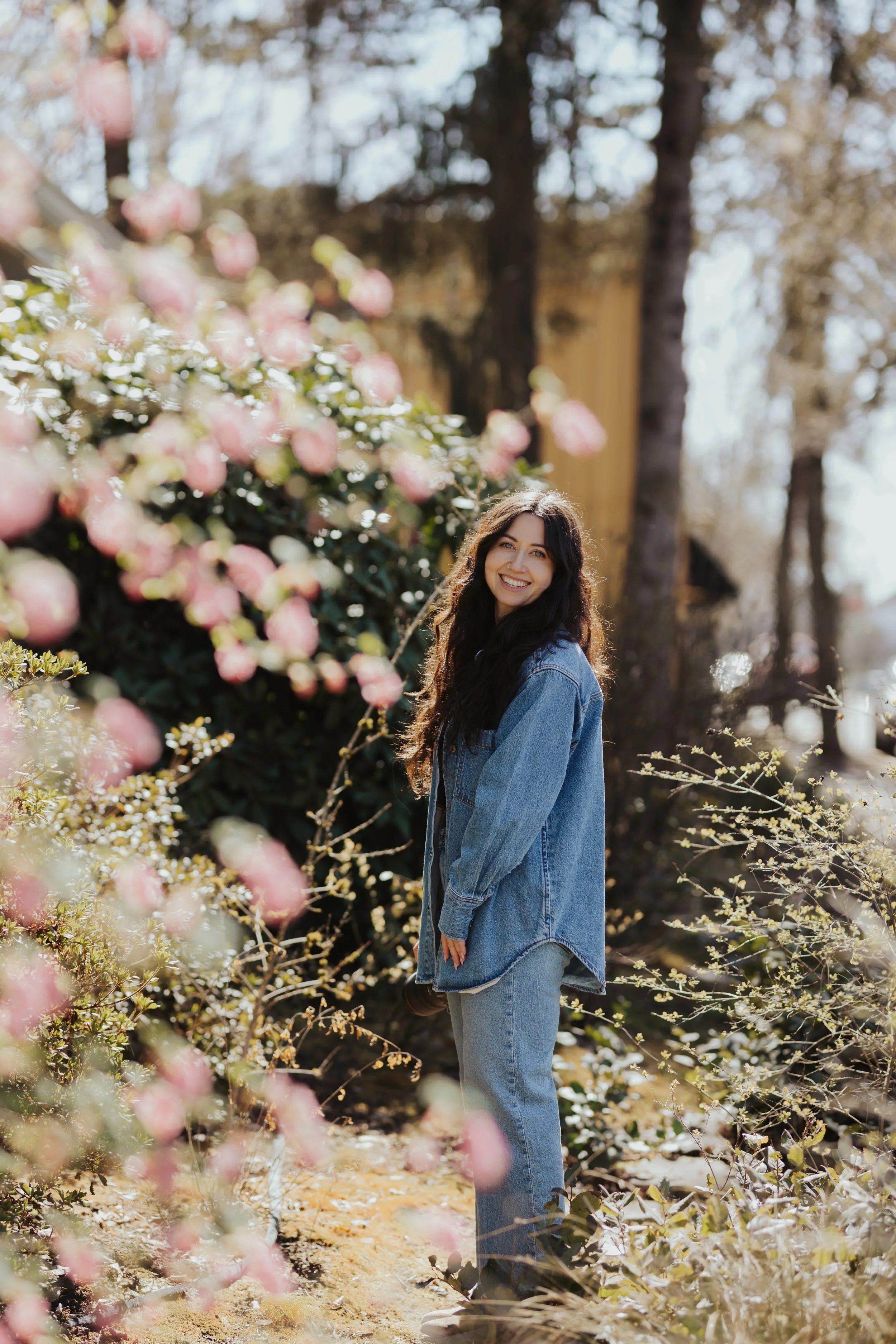 A young woman outdoors in a garden with pink flowers and trees, wearing a denim jacket and smiling.