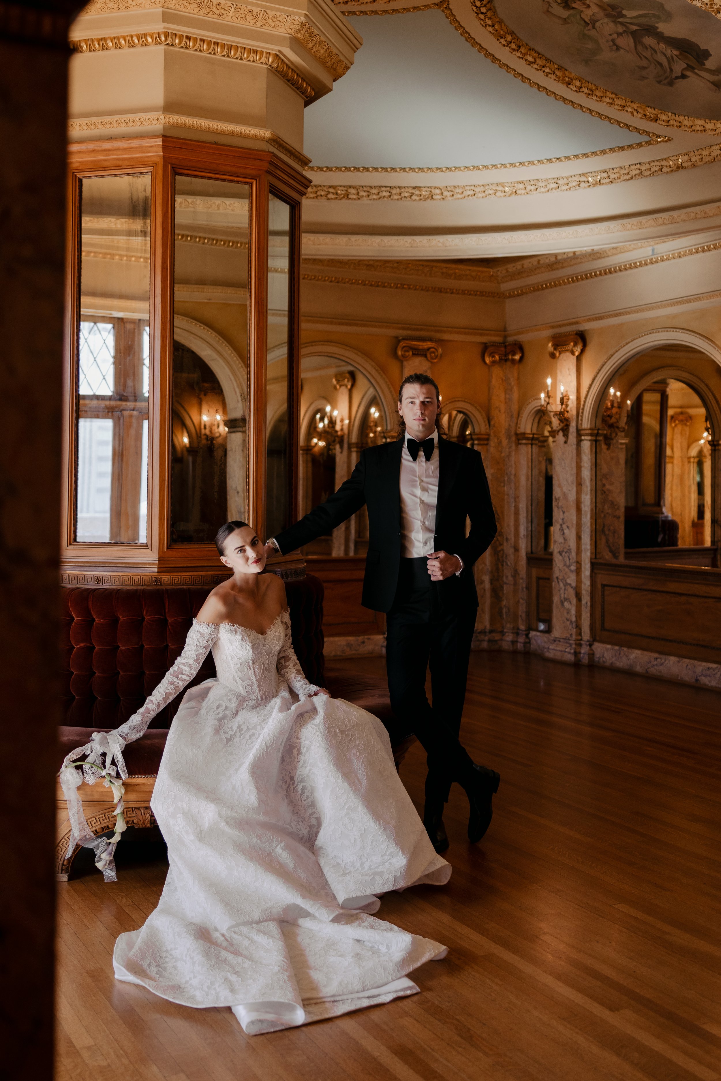 A bride in a white lace wedding dress with off-the-shoulder long sleeves sits on a vintage sofa, holding a small bouquet of flowers with ribbons. A groom in a black tuxedo with a bow tie stands next to her, resting his arm on her shoulder in an opulent room with ornate gold detailing, marble columns, and wooden floors.
