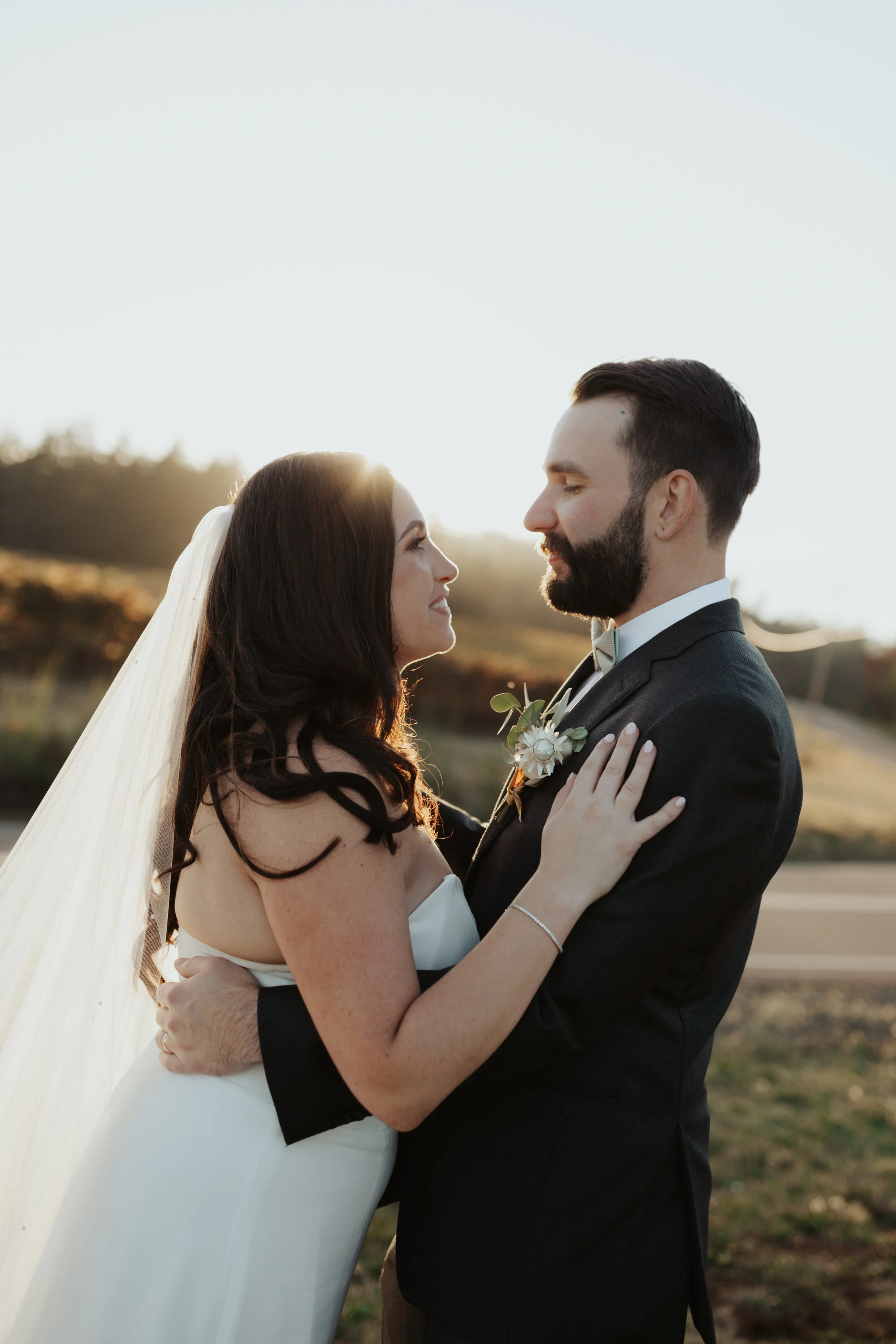 A bride and groom embrace outdoors at sunset, facing each other with the bride's hands on the groom's shoulders and the groom's arms around her waist.